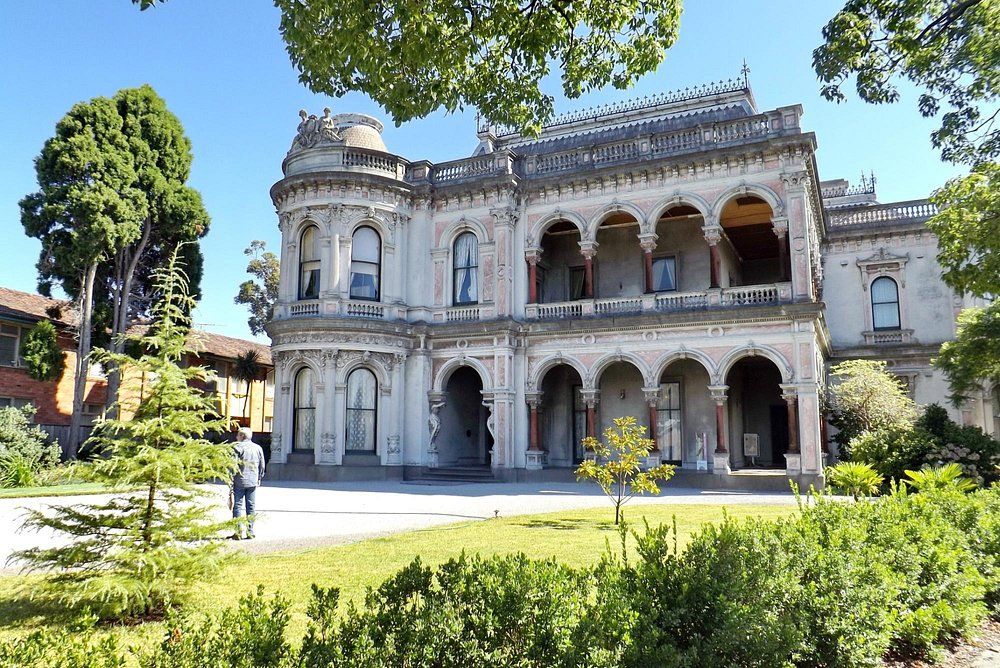Historic Victorian mansion with white stone facade, arched windows and verandah surrounded by manicured gardens in daylight.