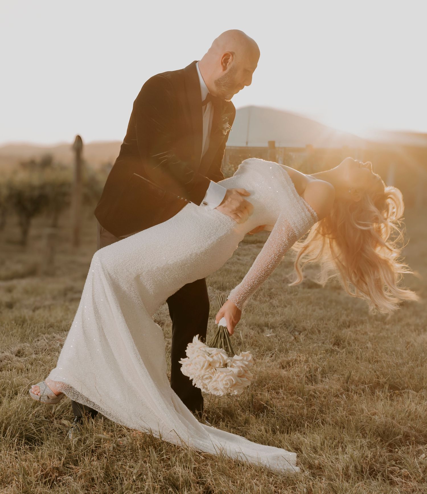 Romantic wedding couple dips in golden sunlit field while bride holds white bouquet in flowing dress.