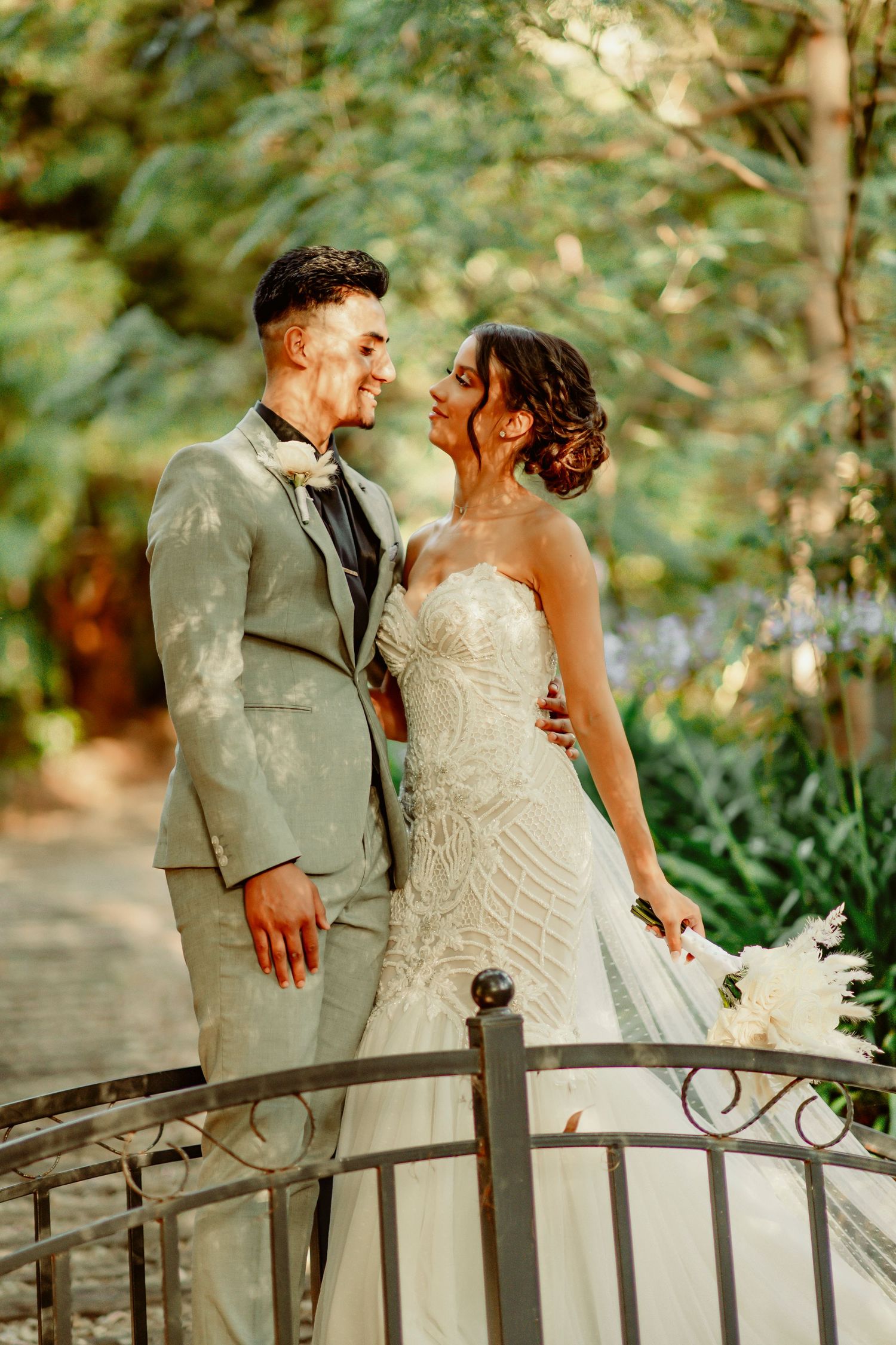 A wedding couple share a joyful moment while standing on a decorative bridge in a lush garden setting.