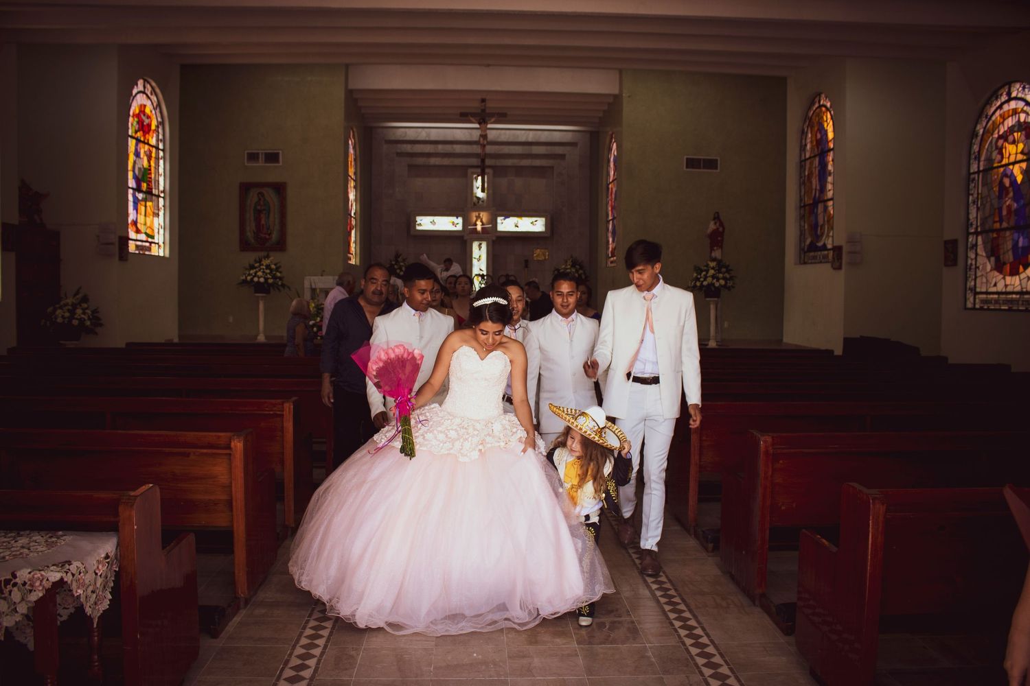 A wedding ceremony in a church with stained glass windows and wooden pews as the wedding party walks down the aisle.