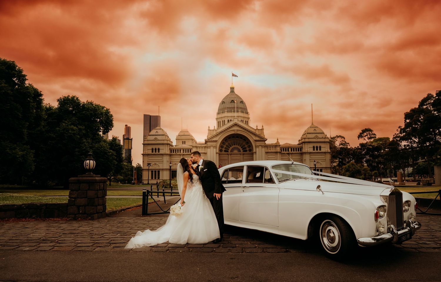 A vintage white Rolls-Royce parked in front of the Royal Exhibition Building in Melbourne under a dramatic orange sky.