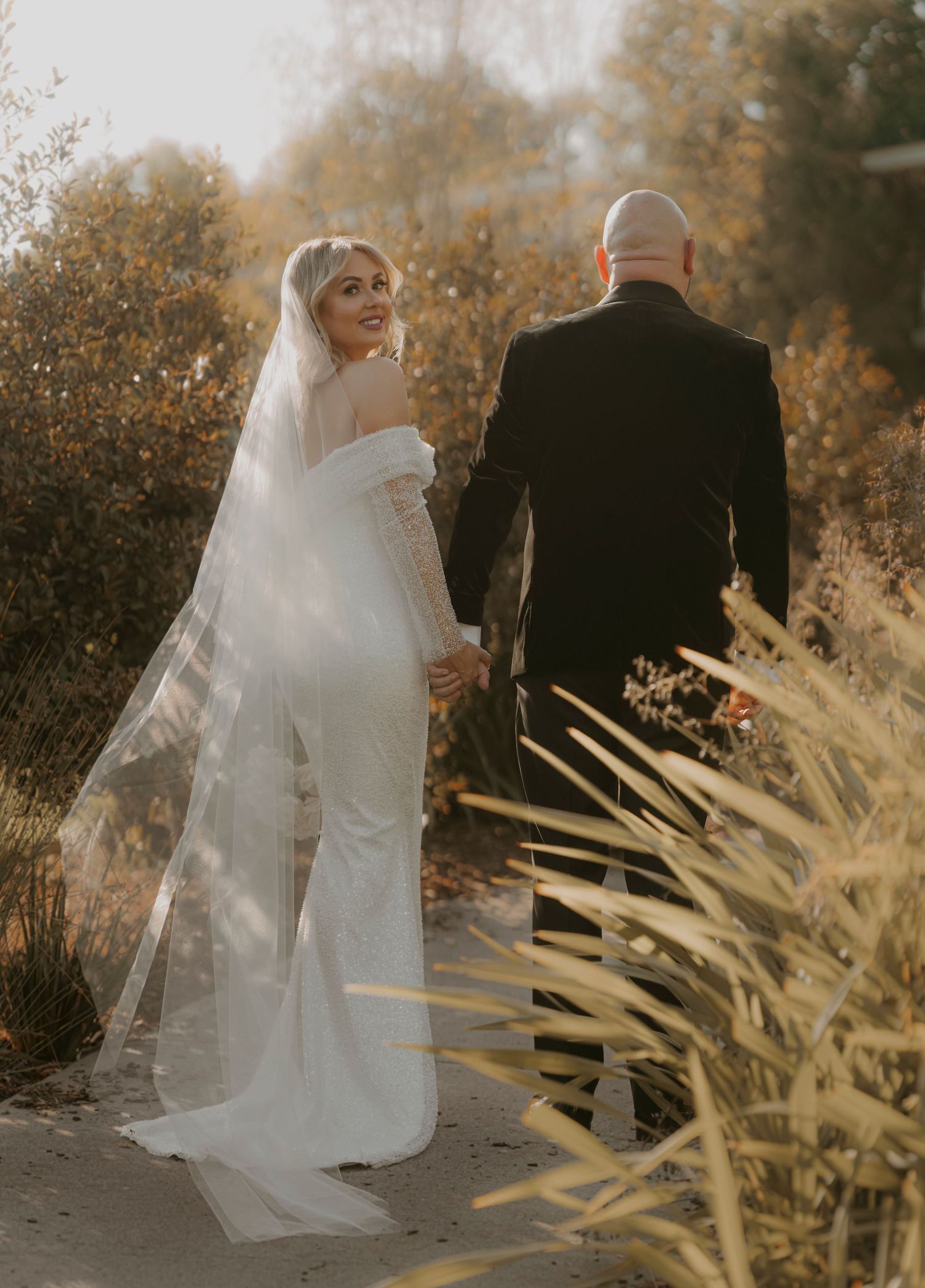 A bride in a white gown and veil glances back while walking with groom on path surrounded by autumn foliage.