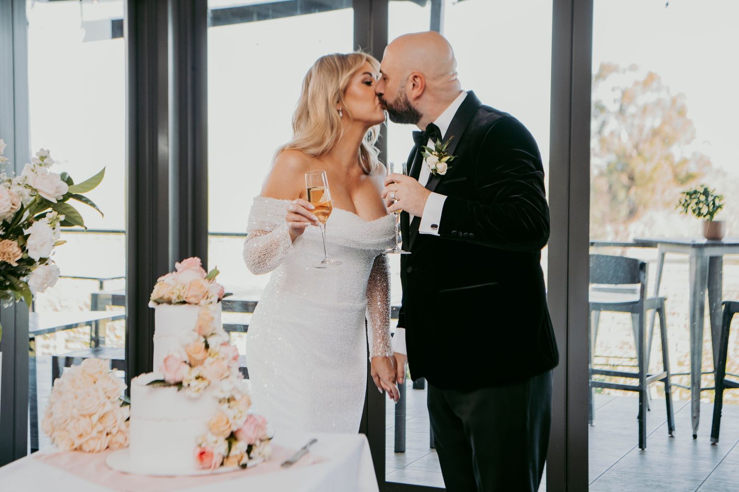 Newlyweds share an intimate moment beside their elegant white and blush wedding cake at a modern reception venue.