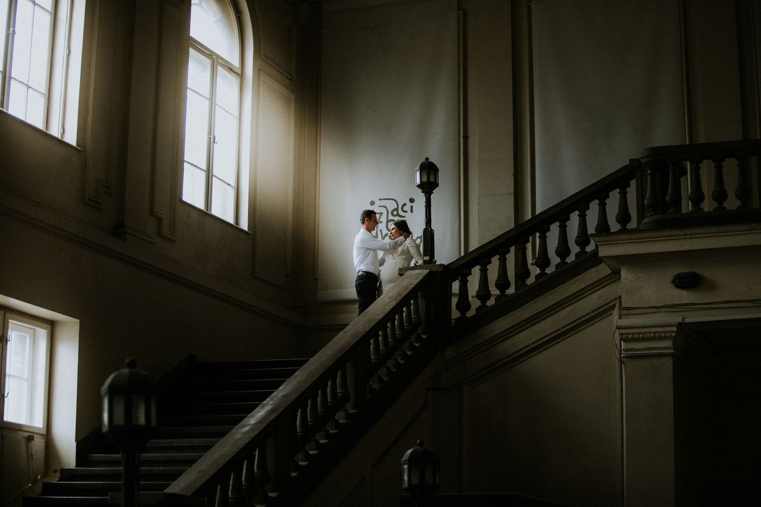 A couple in wedding attire stands atop a grand staircase in a historic building with dramatic lighting through tall windows.
