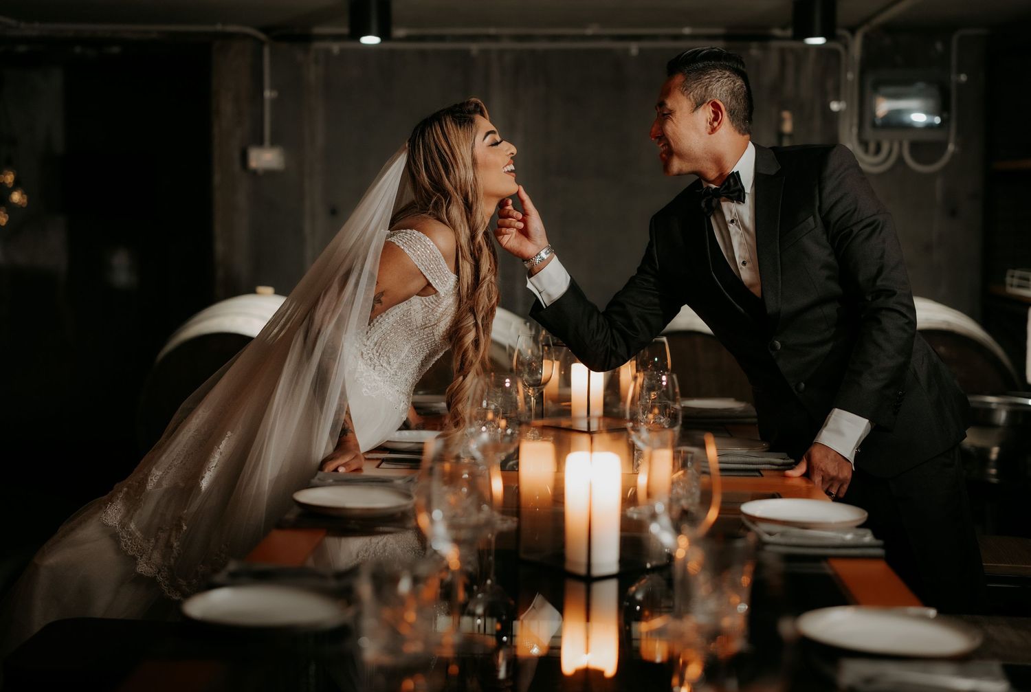 A romantic wedding couple shares an intimate moment at a candlelit dining table in a dark, moody restaurant setting.
