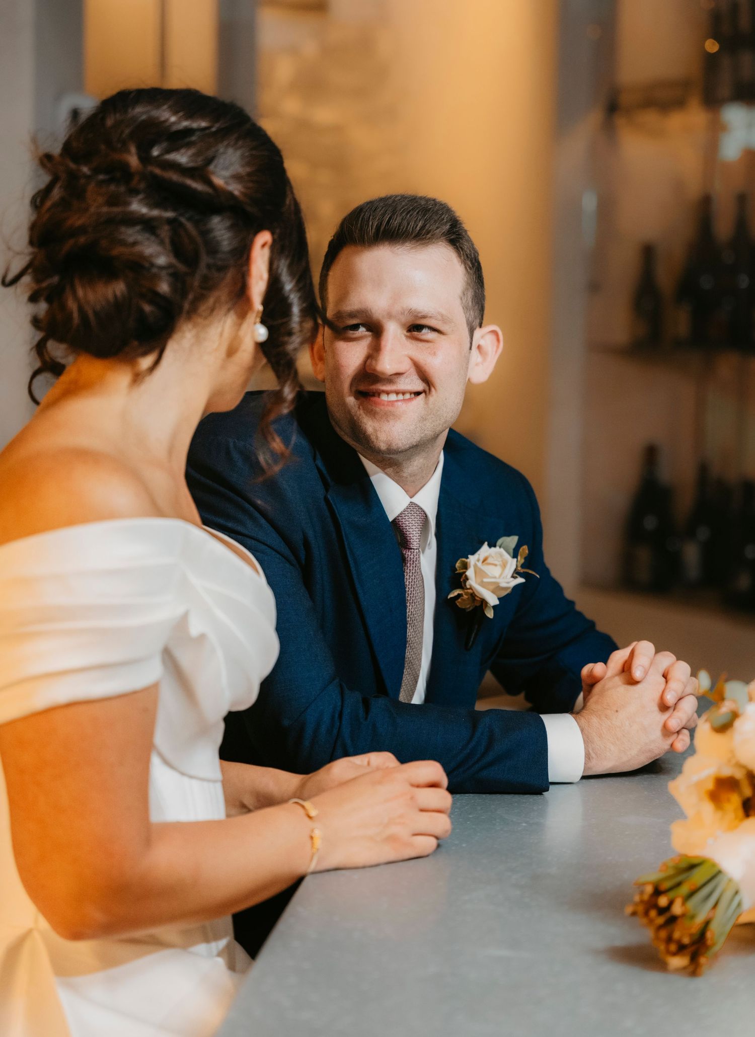 Happy couple sitting at table together on their wedding day wearing bridal gown and navy suit with boutonniere.
