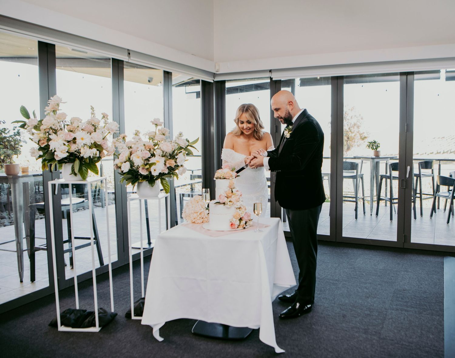 A newlywed couple cuts their wedding cake together at an elegant reception venue with floral arrangements on pedestals.