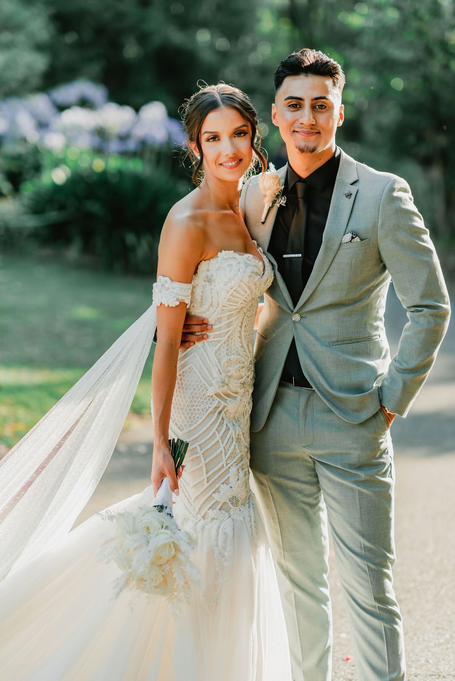 A bride in an off-shoulder lace gown and a groom in a light gray suit pose together outdoors for a wedding photo.