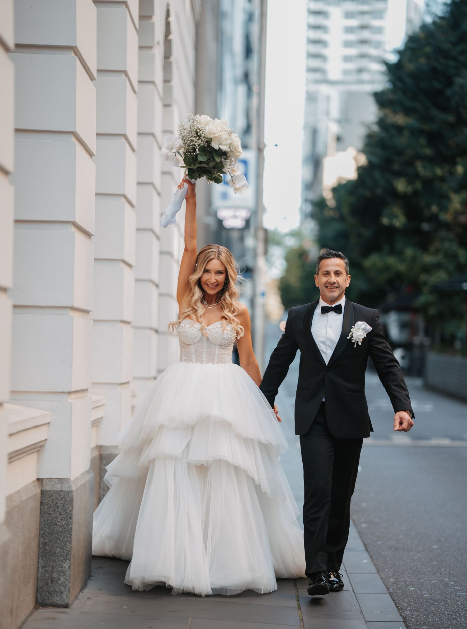 A newlywed couple walks along a city sidewalk as the bride joyfully raises her bouquet in celebration.