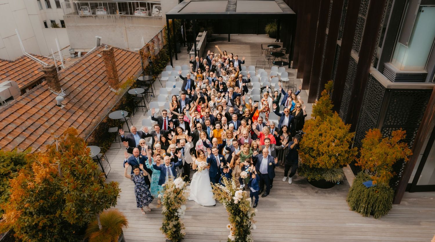 Wedding guests gather for an aerial photo on an outdoor terrace surrounded by fall foliage and modern architecture.