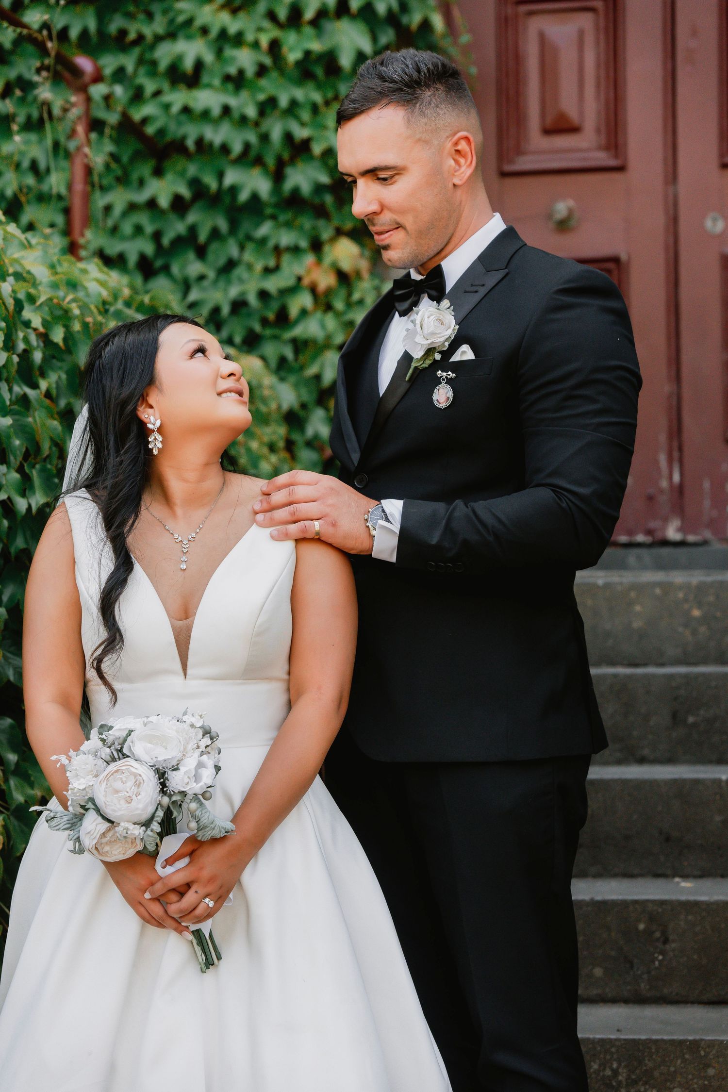 A romantic wedding couple poses on stone steps with ivy-covered walls and a rustic door behind them.