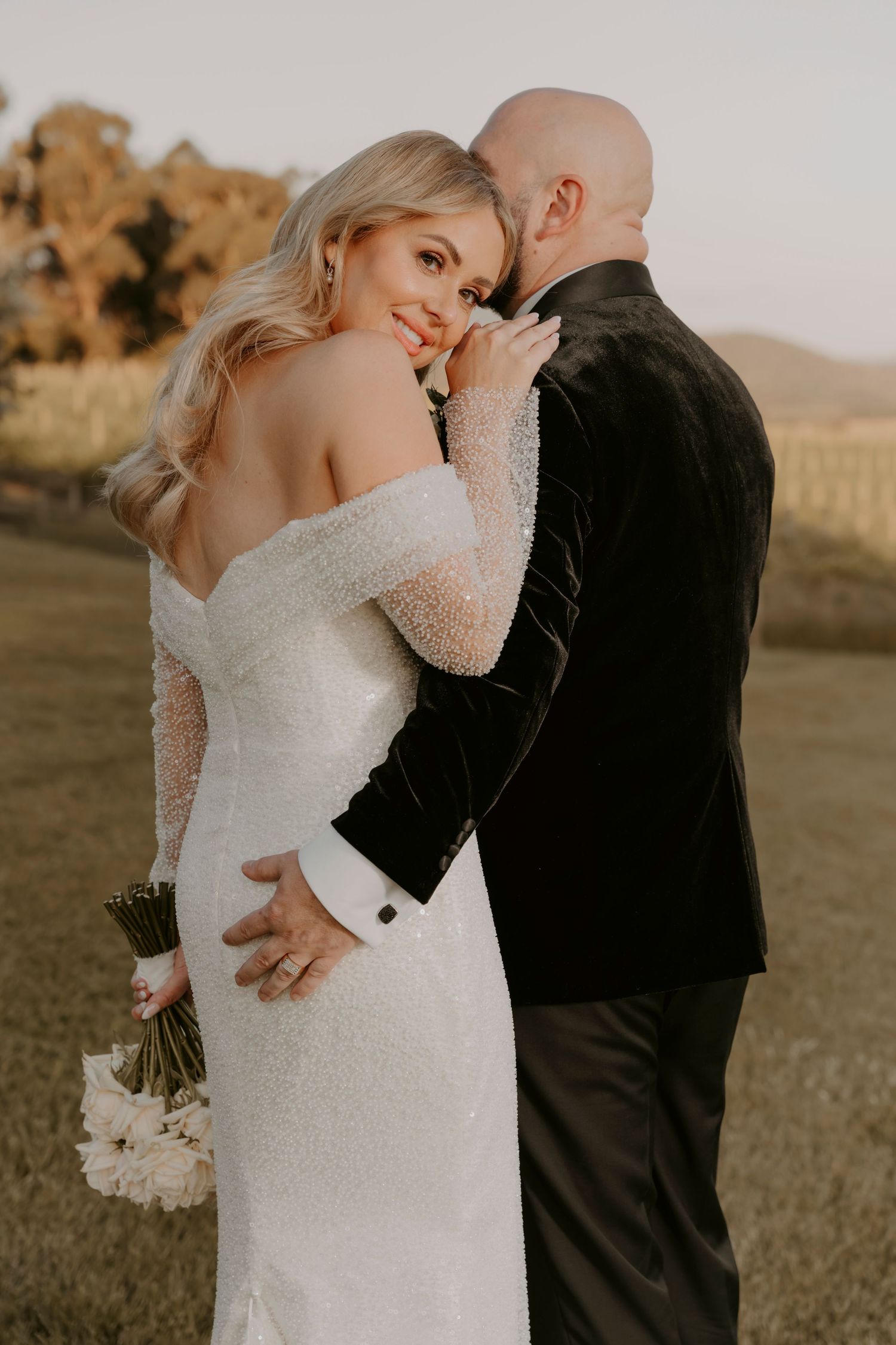 A romantic wedding couple embraces at sunset, with the bride in a fitted white off-shoulder gown and groom in black suit.
