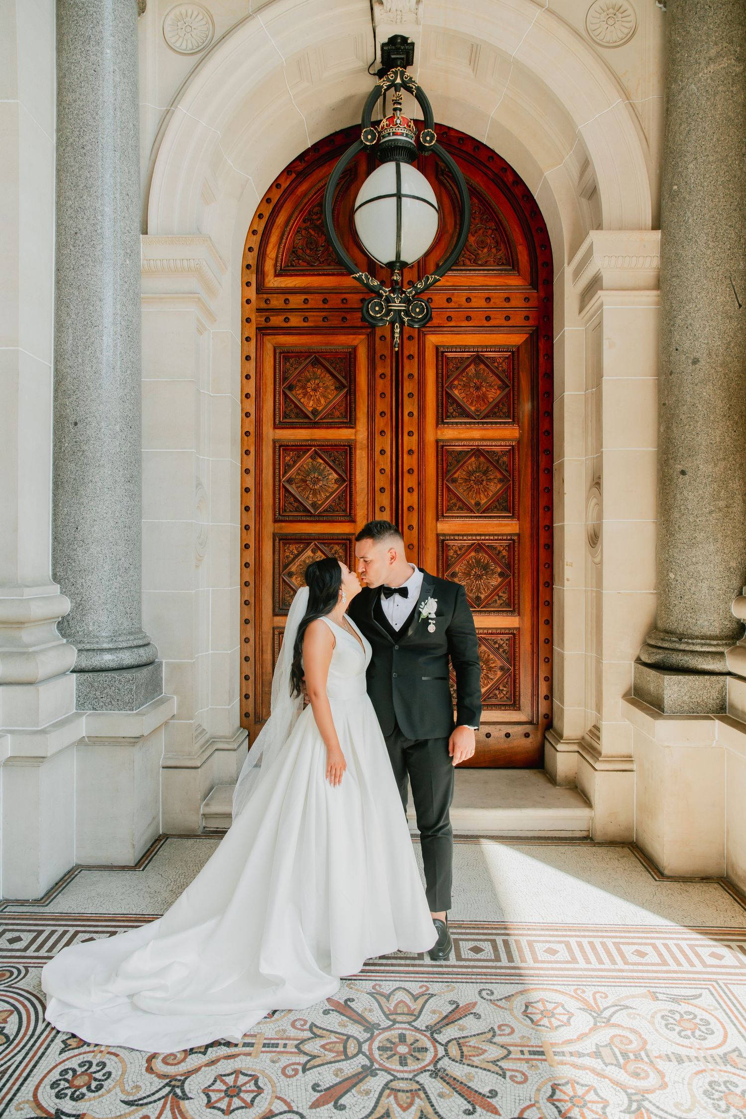 A couple poses beneath an ornate archway with detailed wooden doors and tiled flooring at their wedding.