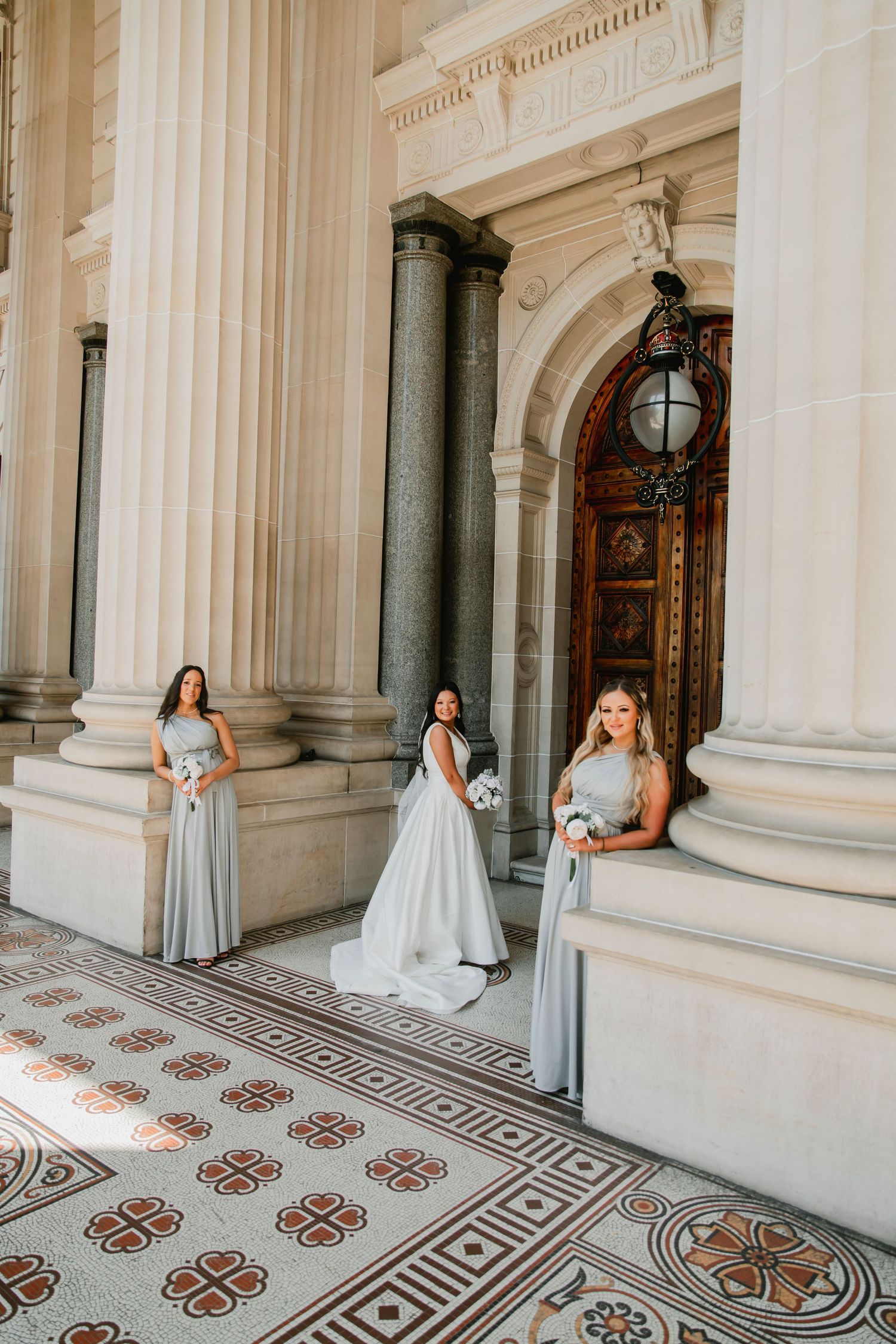 Three people in formal attire stand between elegant marble columns on ornate tiled floor at historic building entrance.