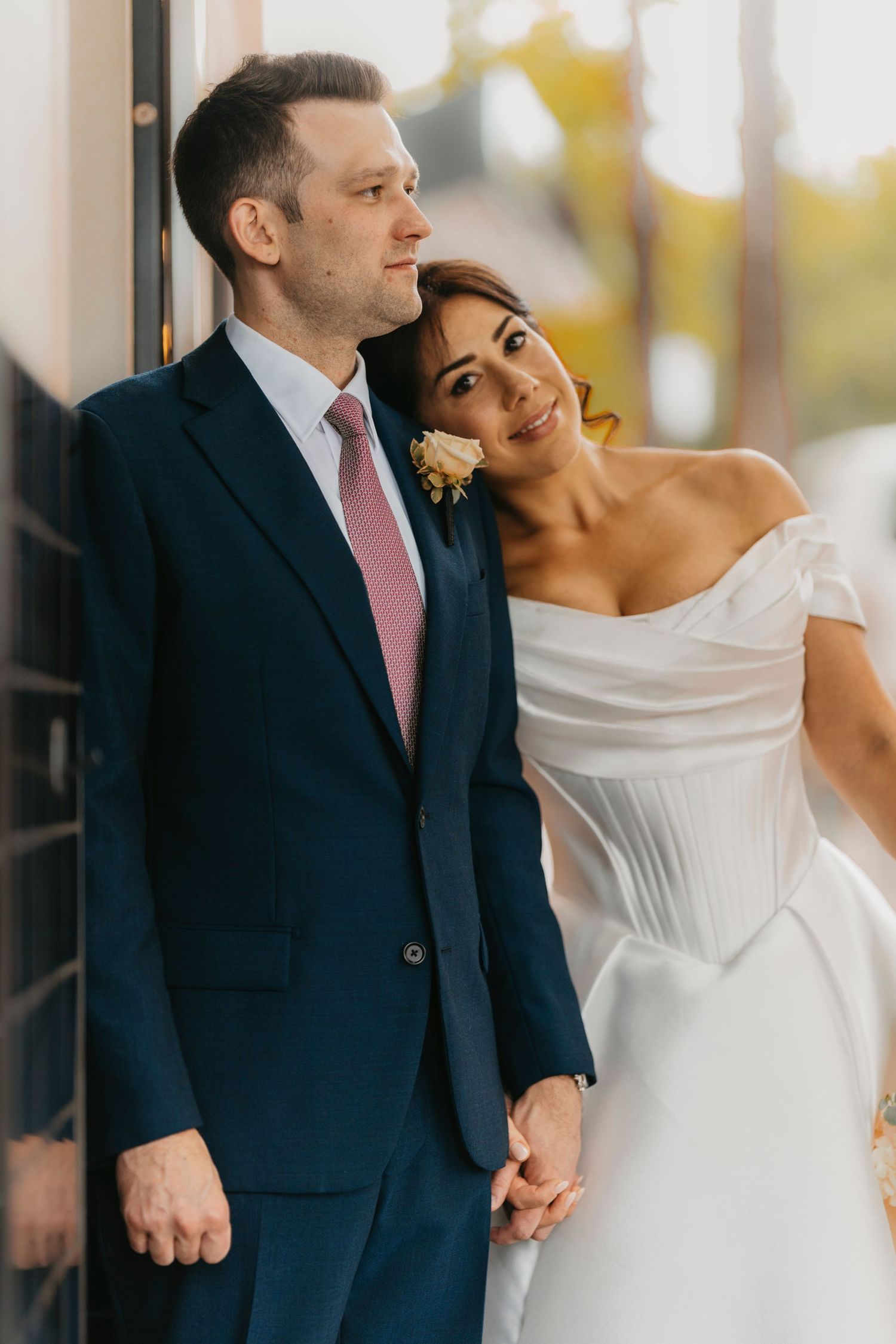 Elegant wedding couple in navy suit and off-shoulder white dress pose together against natural outdoor background.