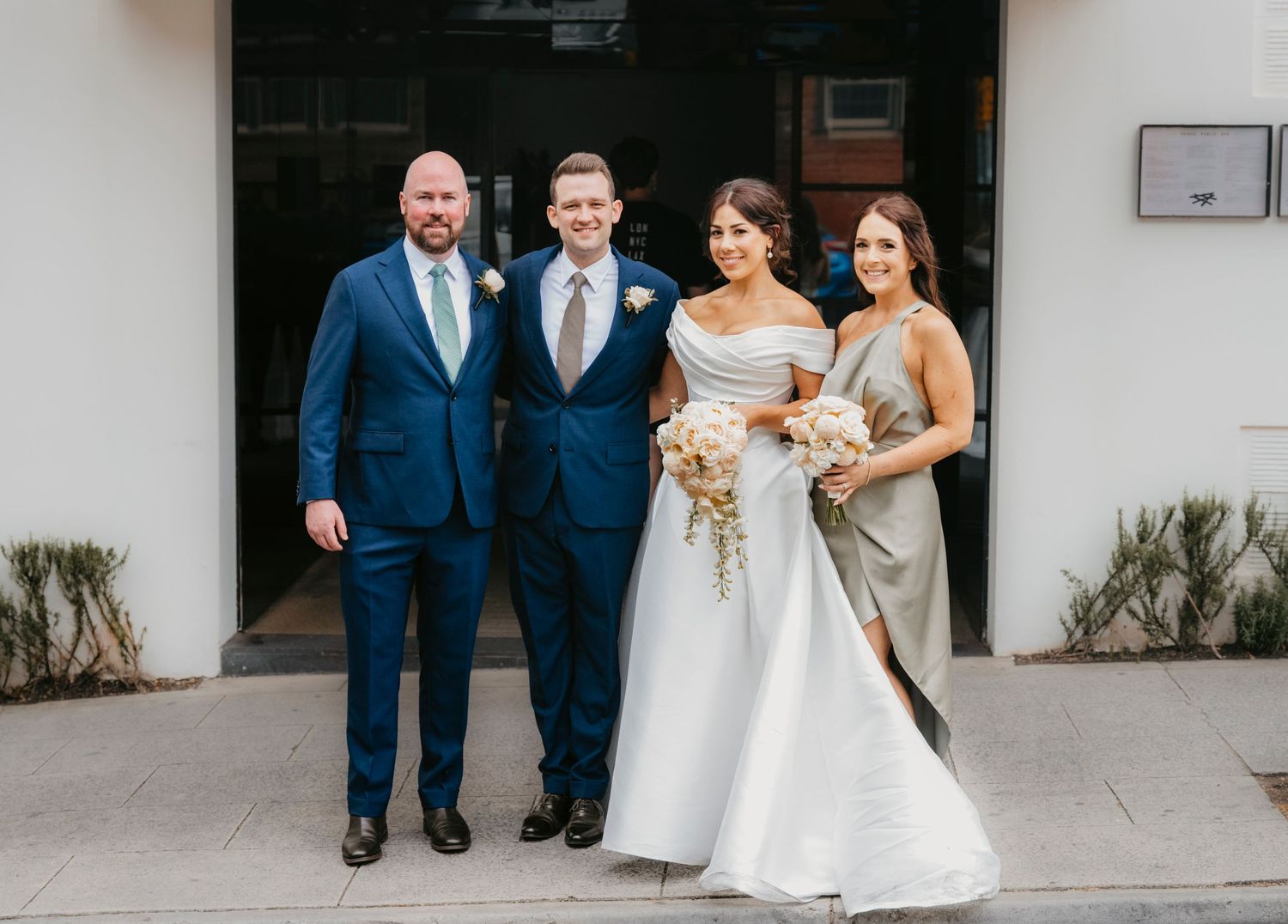 Wedding party of four stands together outside venue entrance, with bride in white gown holding floral bouquet.