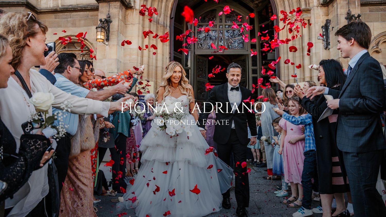 Wedding guests toss red heart-shaped confetti as newlyweds exit ceremony beneath vintage brick archway.
