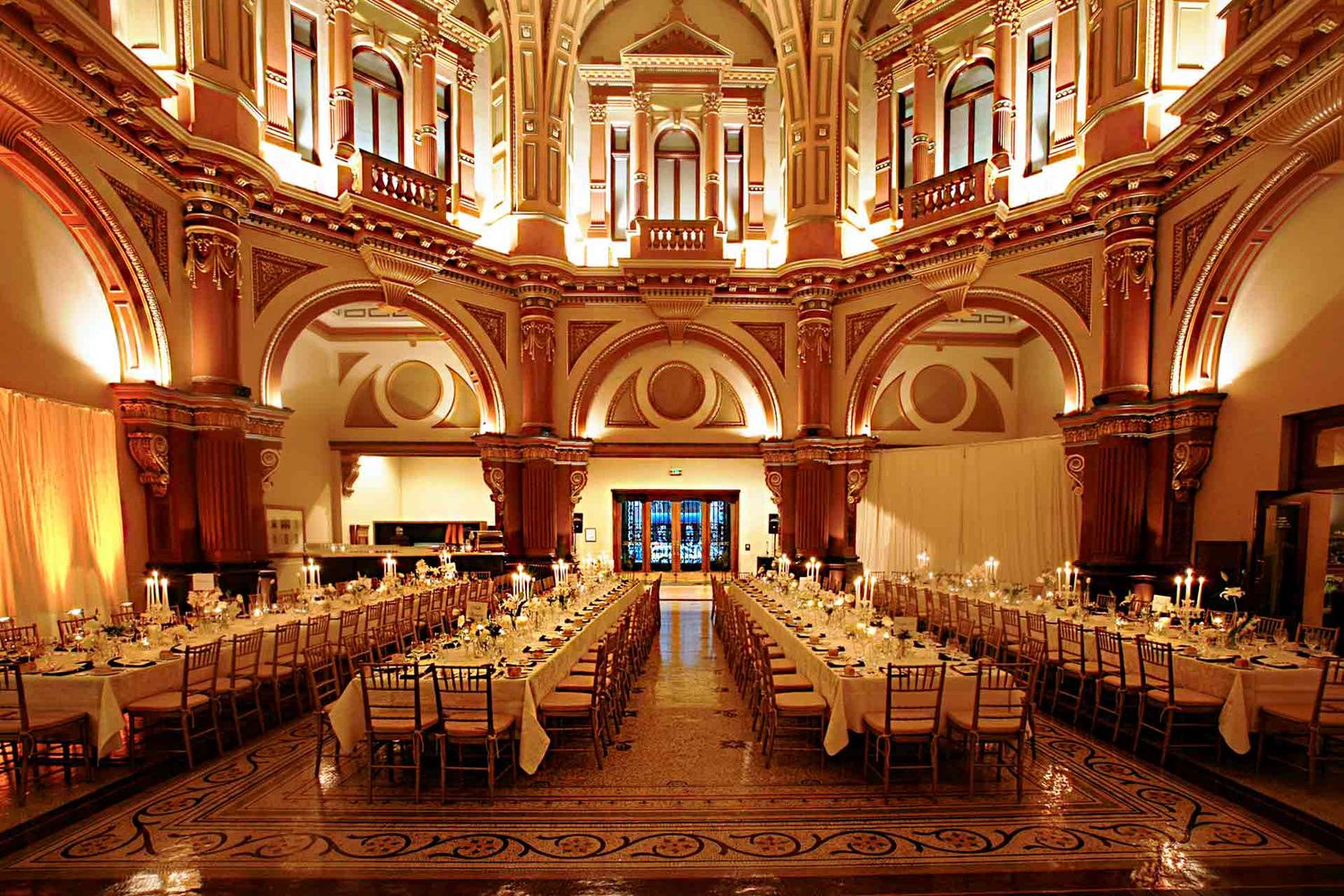 Elegant banquet hall with ornate architecture and golden lighting features long dining tables arranged in rows on polished floors.