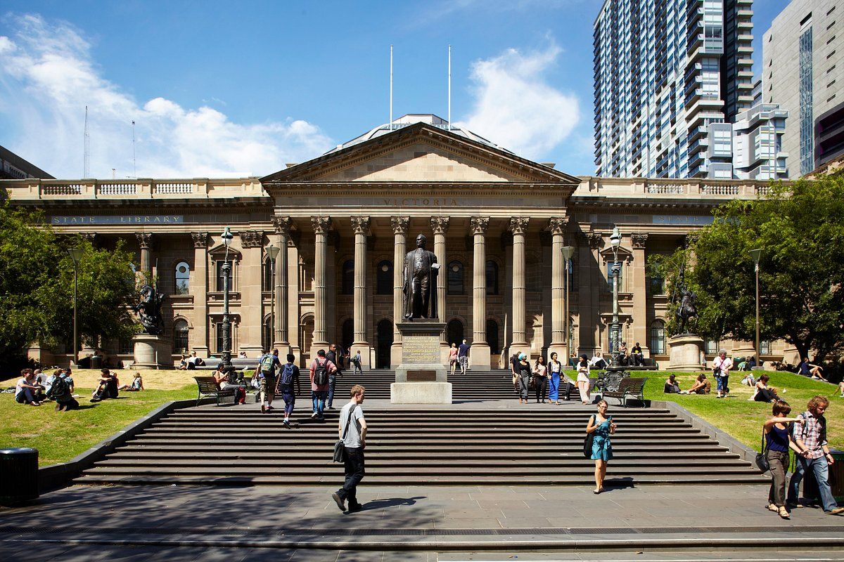 The neoclassical State Library of Victoria building with grand columns and steps where visitors gather on a sunny day in Melbourne.