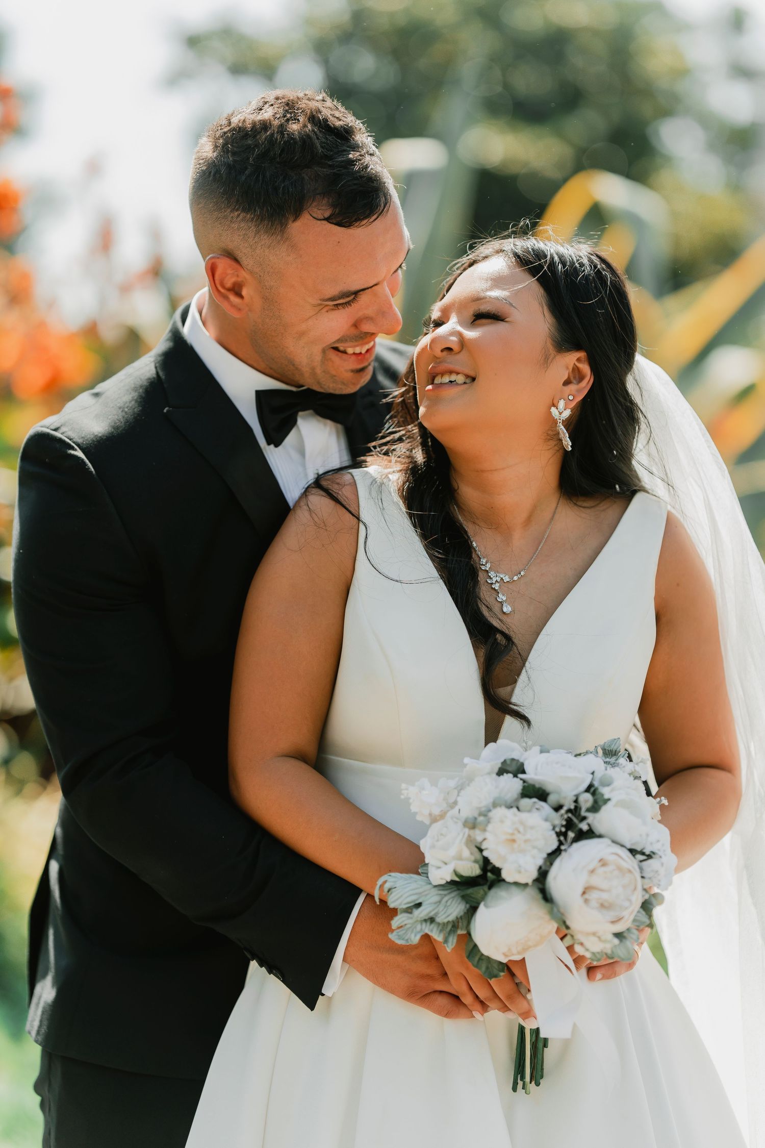 A joyful wedding couple share an intimate moment outdoors while the bride holds a white rose bouquet.