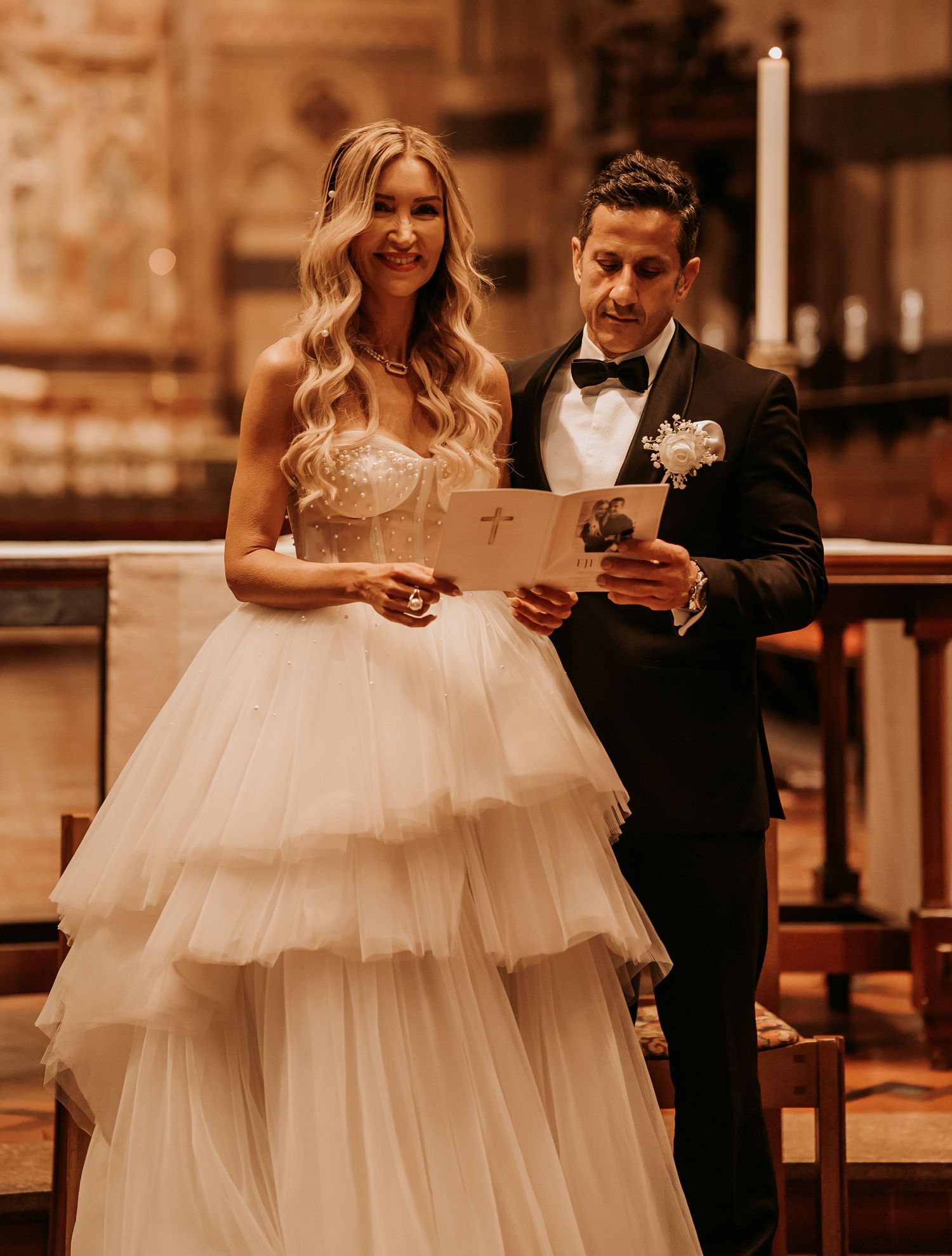A bride in a layered tulle gown and groom in classic tuxedo read from papers during a wedding ceremony in church.