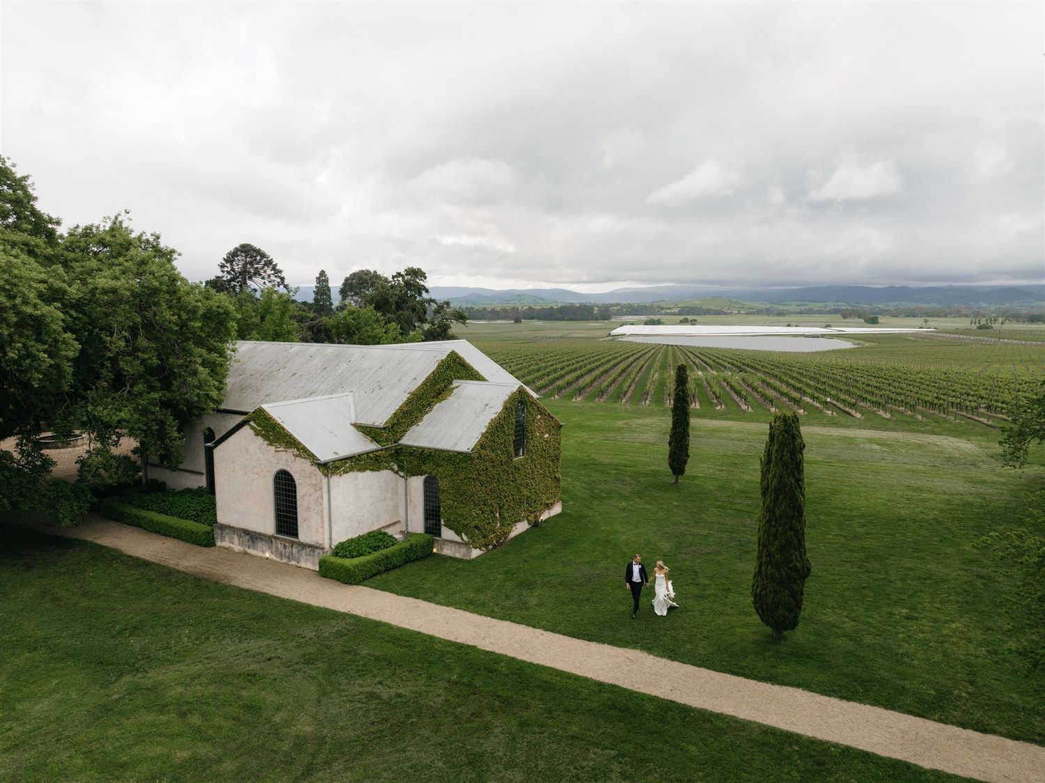 Aerial view of a white chapel surrounded by lush vineyards and cypress trees with distant rolling hills and water views.