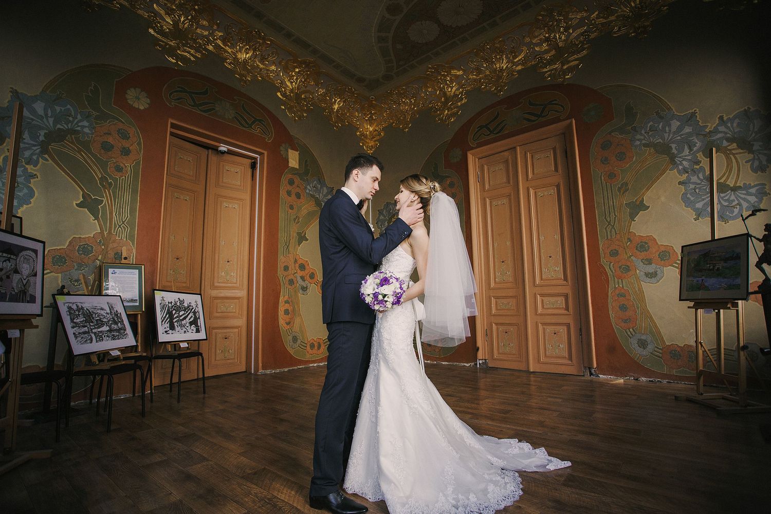 Elegant wedding couple embraces in ornate historic room with painted walls and wood floors.