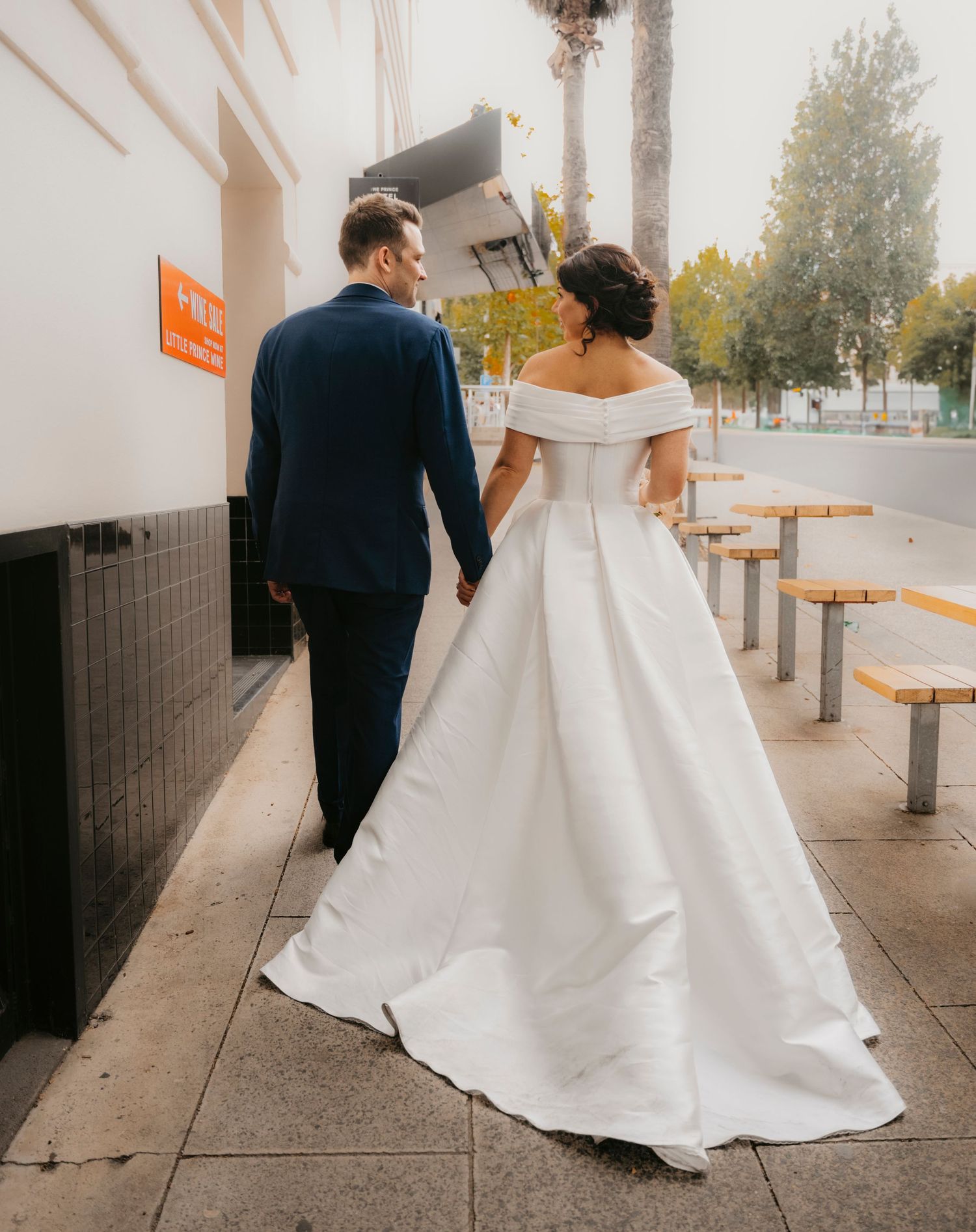 A couple in wedding attire walk hand in hand down a sidewalk with their backs to the camera and trees in the distance.
