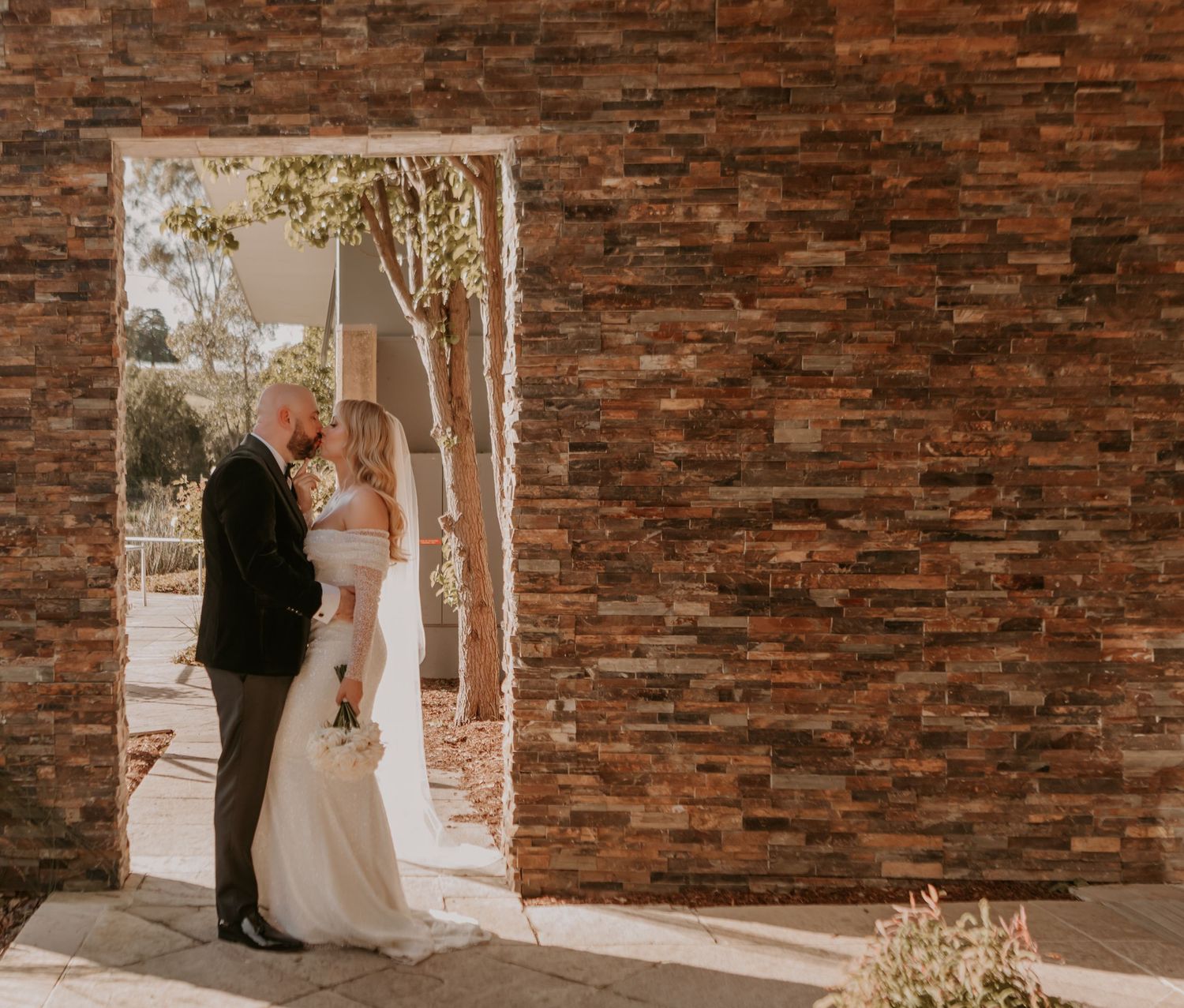 Newlyweds share an intimate moment framed by rustic brick walls and sunlit birch trees on their wedding day.