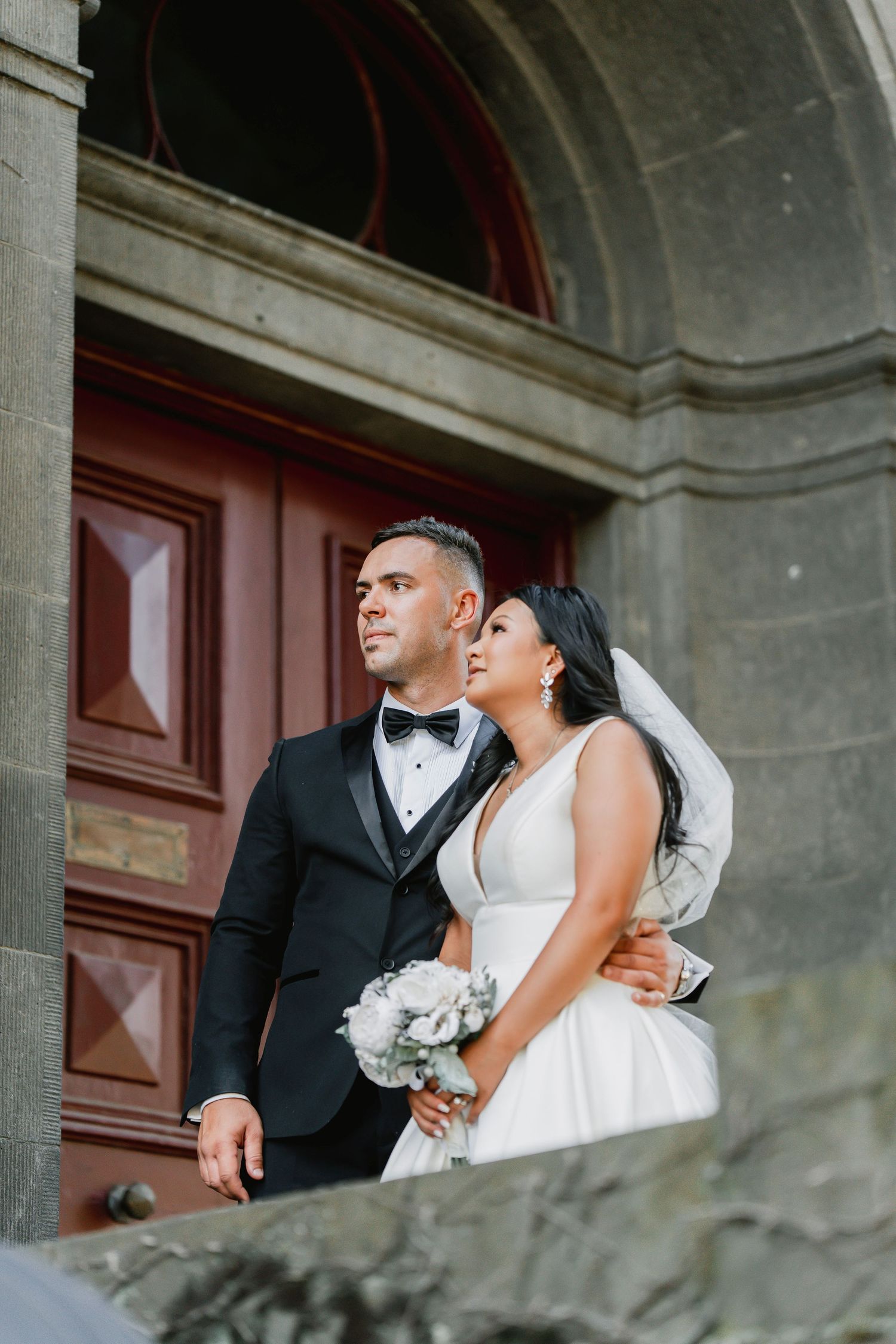 A couple in elegant wedding attire stand together in a grand stone archway entrance with wooden doors.