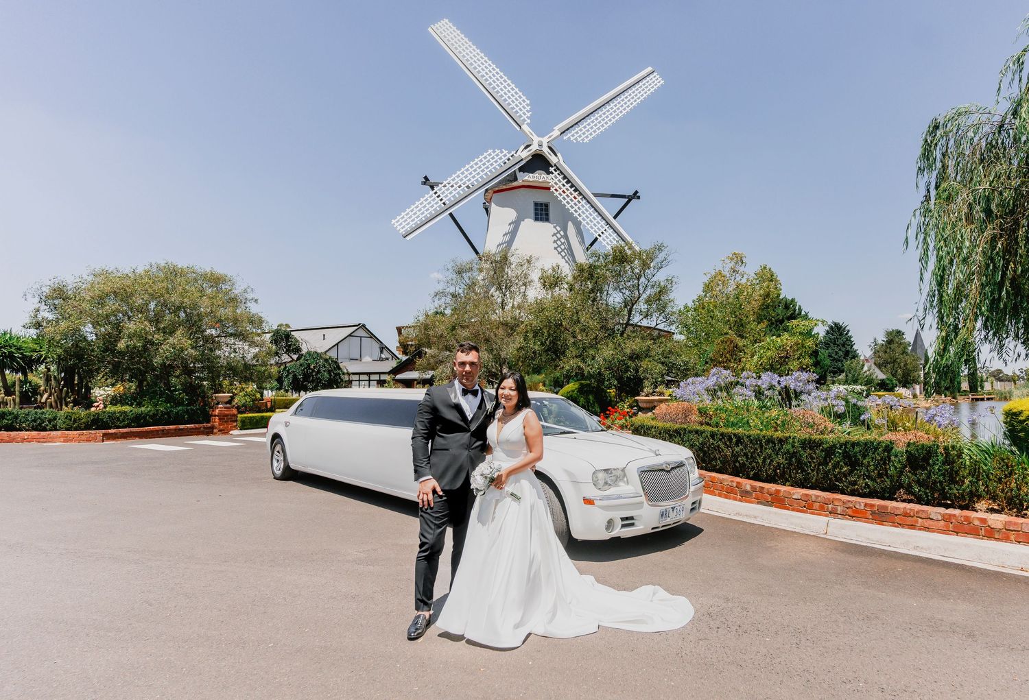 A white limousine parked in front of a traditional Dutch windmill with elegant wedding couple posing nearby.