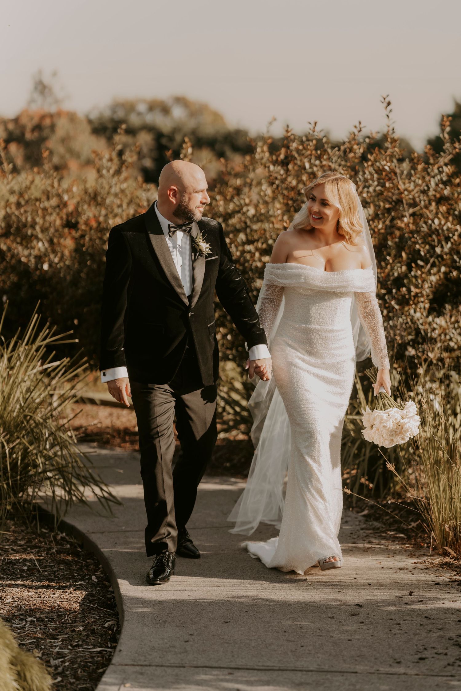 A couple in wedding attire walks together on a garden path surrounded by autumn foliage at sunset.