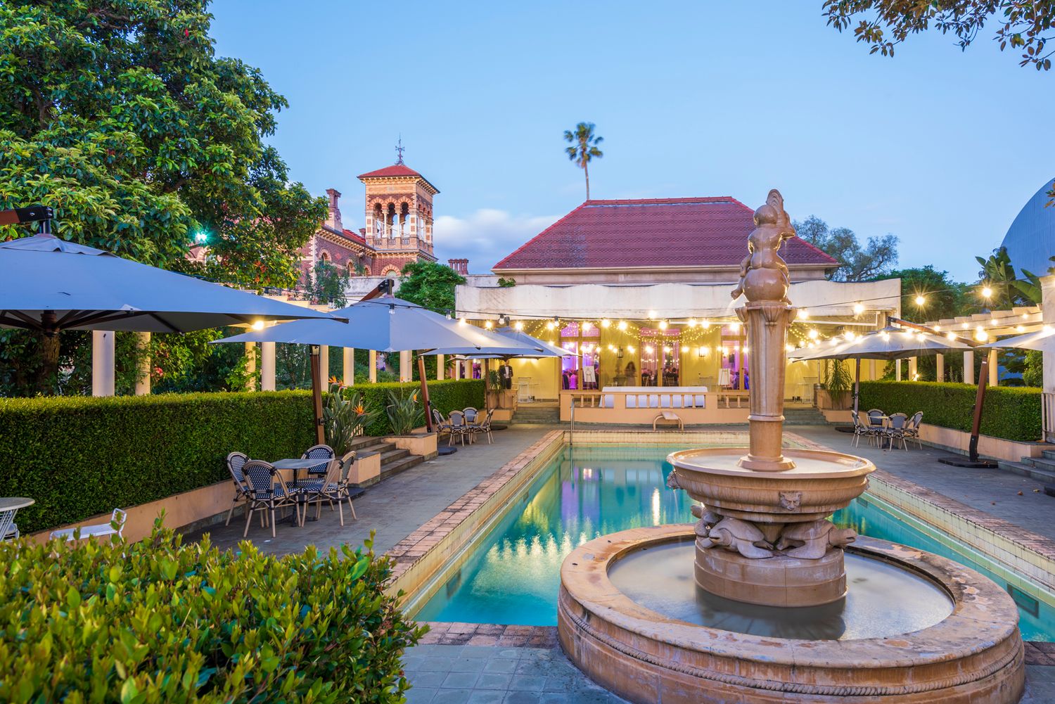 Luxurious hotel courtyard with illuminated swimming pool and ornate fountain at dusk features Mediterranean architecture.