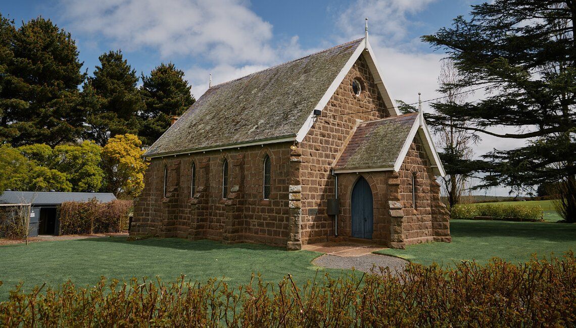 A historic stone church with wooden shingles sits on a grassy landscape under blue skies in rural countryside.