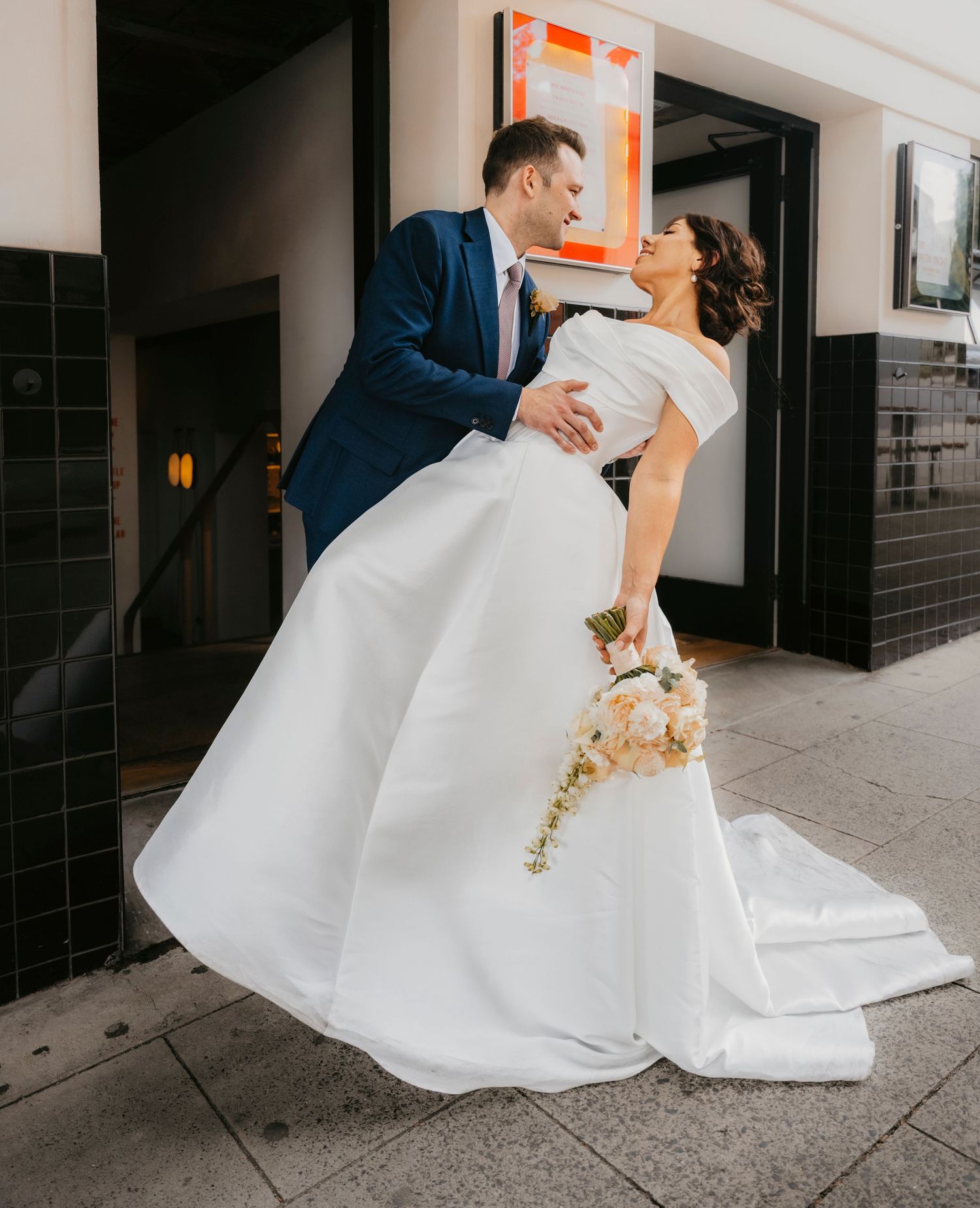 A couple dips romantically in wedding attire on a city sidewalk, with the bride holding a cascading floral bouquet.
