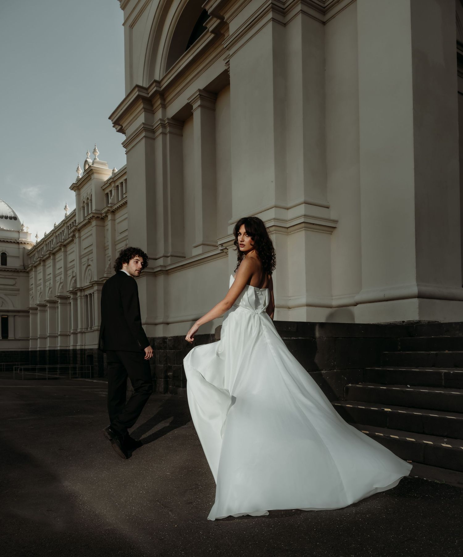 A couple in formal attire poses on grand stone steps outside a historic building with elegant architectural details.