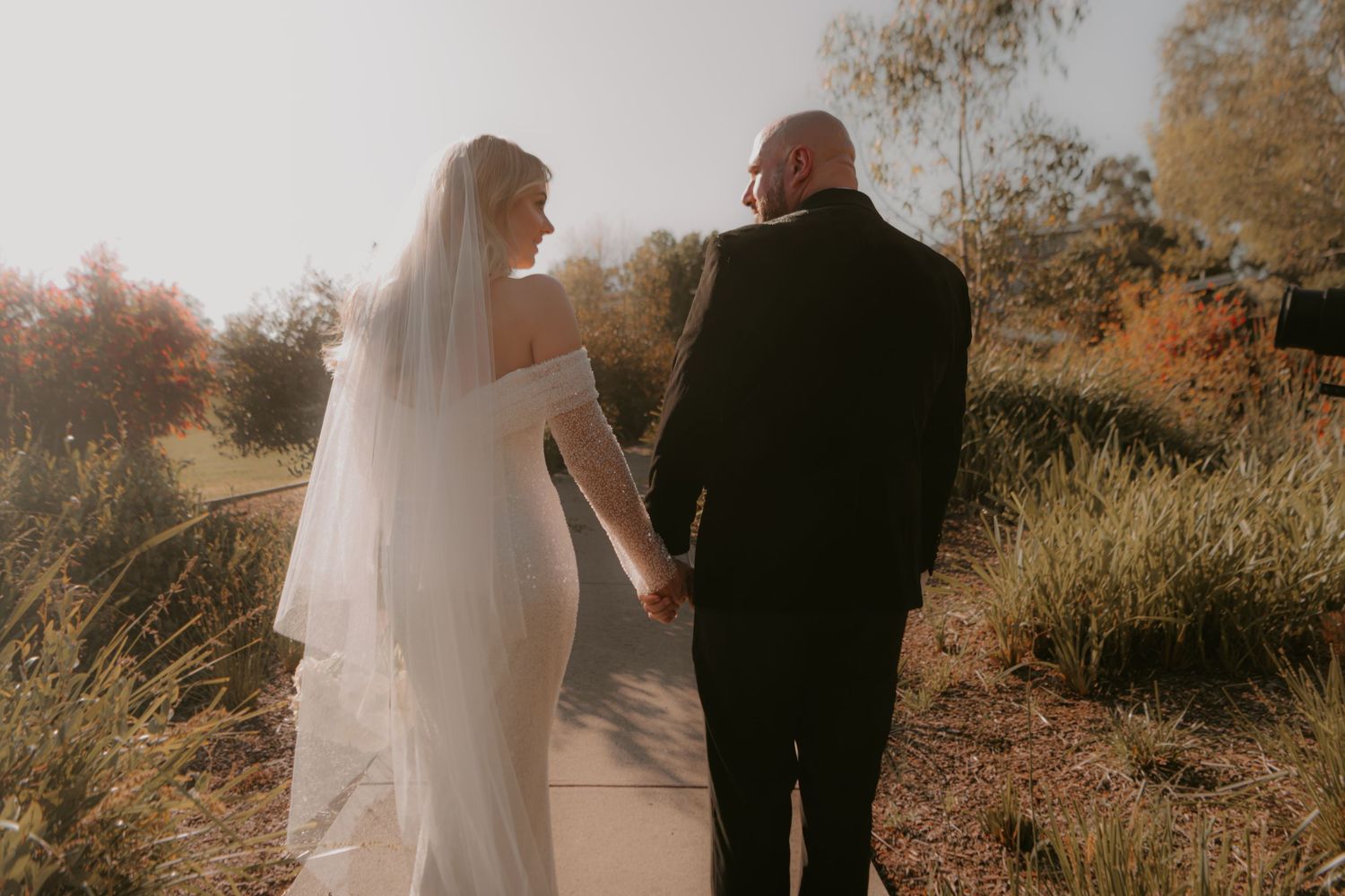 A couple holding hands walking down an outdoor path during golden hour while dressed in wedding attire.