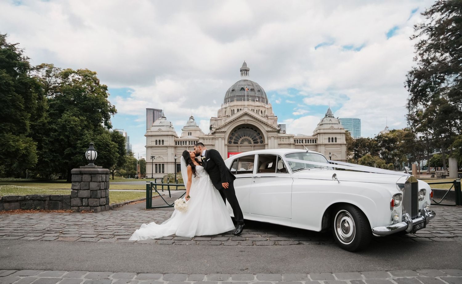 A white vintage Rolls-Royce parked in front of the Royal Exhibition Building in Melbourne with wedding couple beside it.