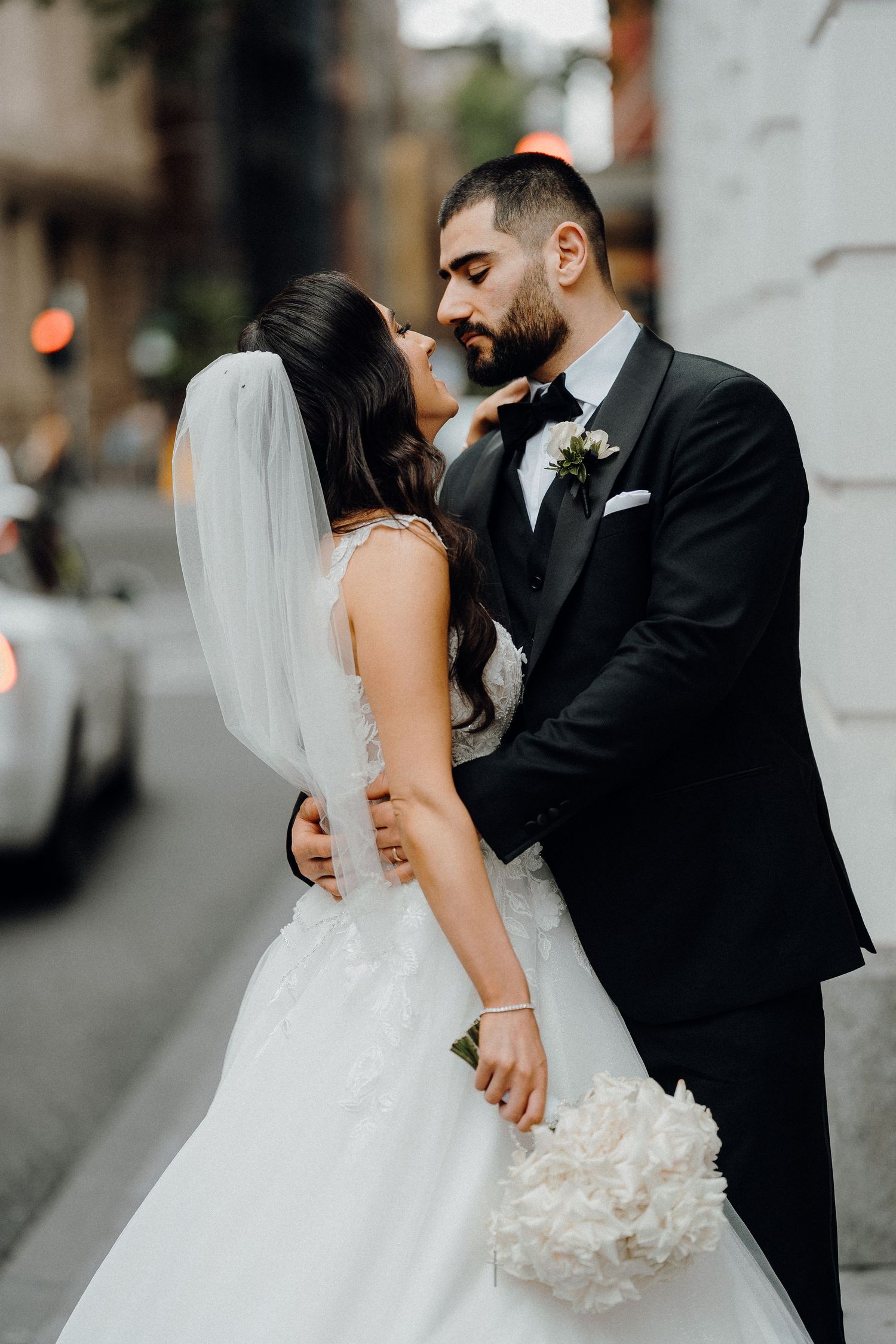 A bride in a white ballgown and veil shares an intimate moment with their spouse on a city street.