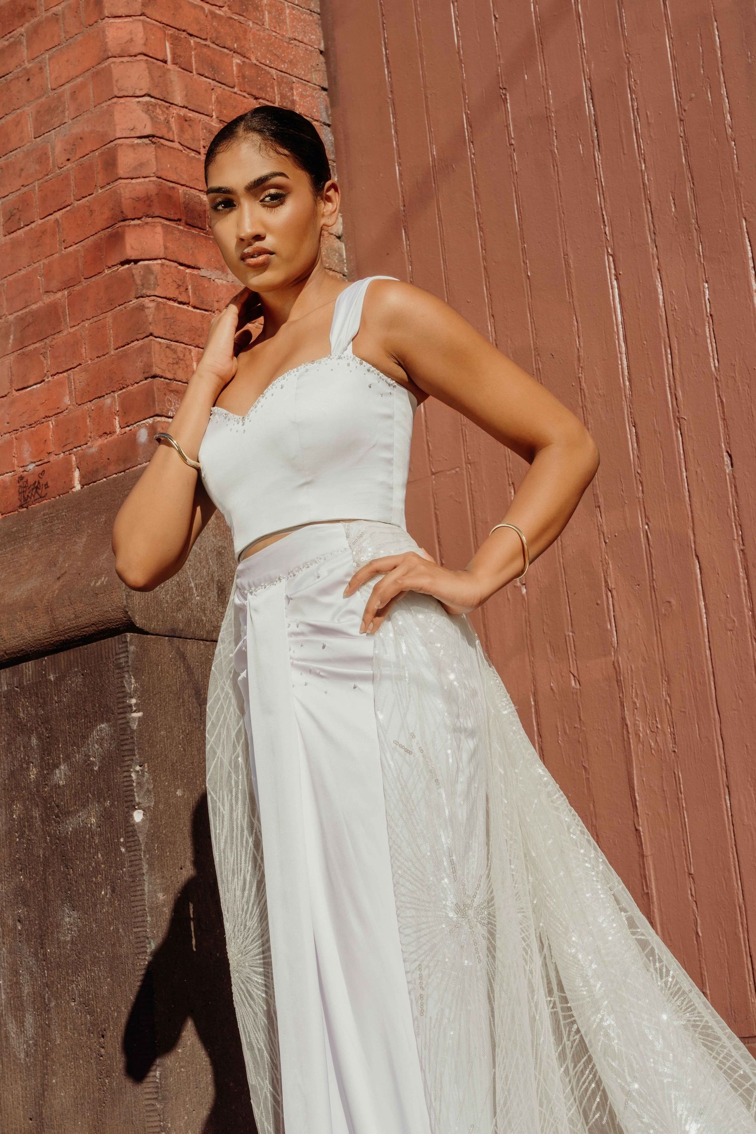 A model poses in a flowing white gown with a fitted bodice against a brick wall in warm sunlight.