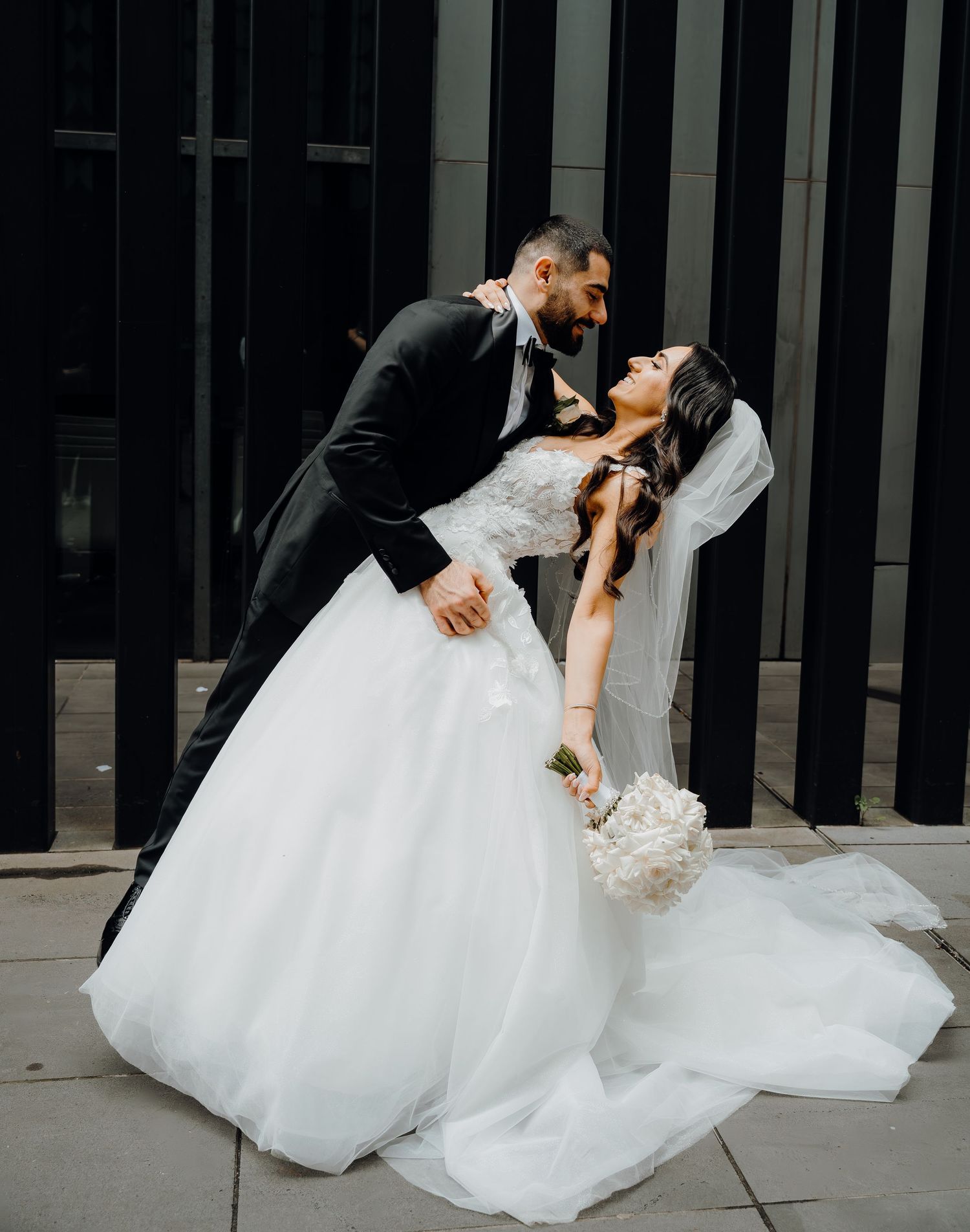 A newlywed couple shares a romantic dip pose in front of a modern building, with the bride's white dress flowing elegantly.