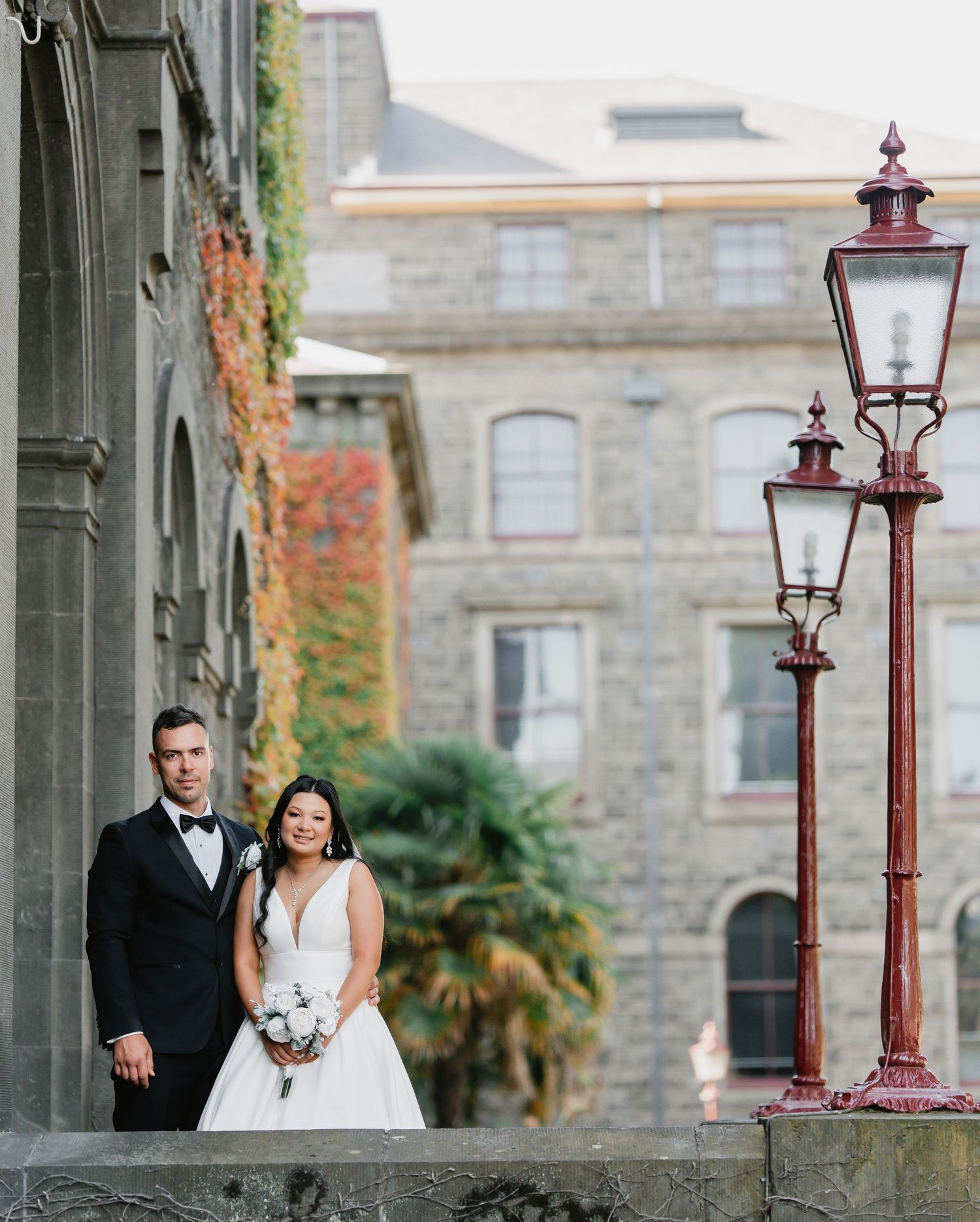 Elegant couple poses by vintage stone building exterior with ornate red lamp post and climbing vines on wall.