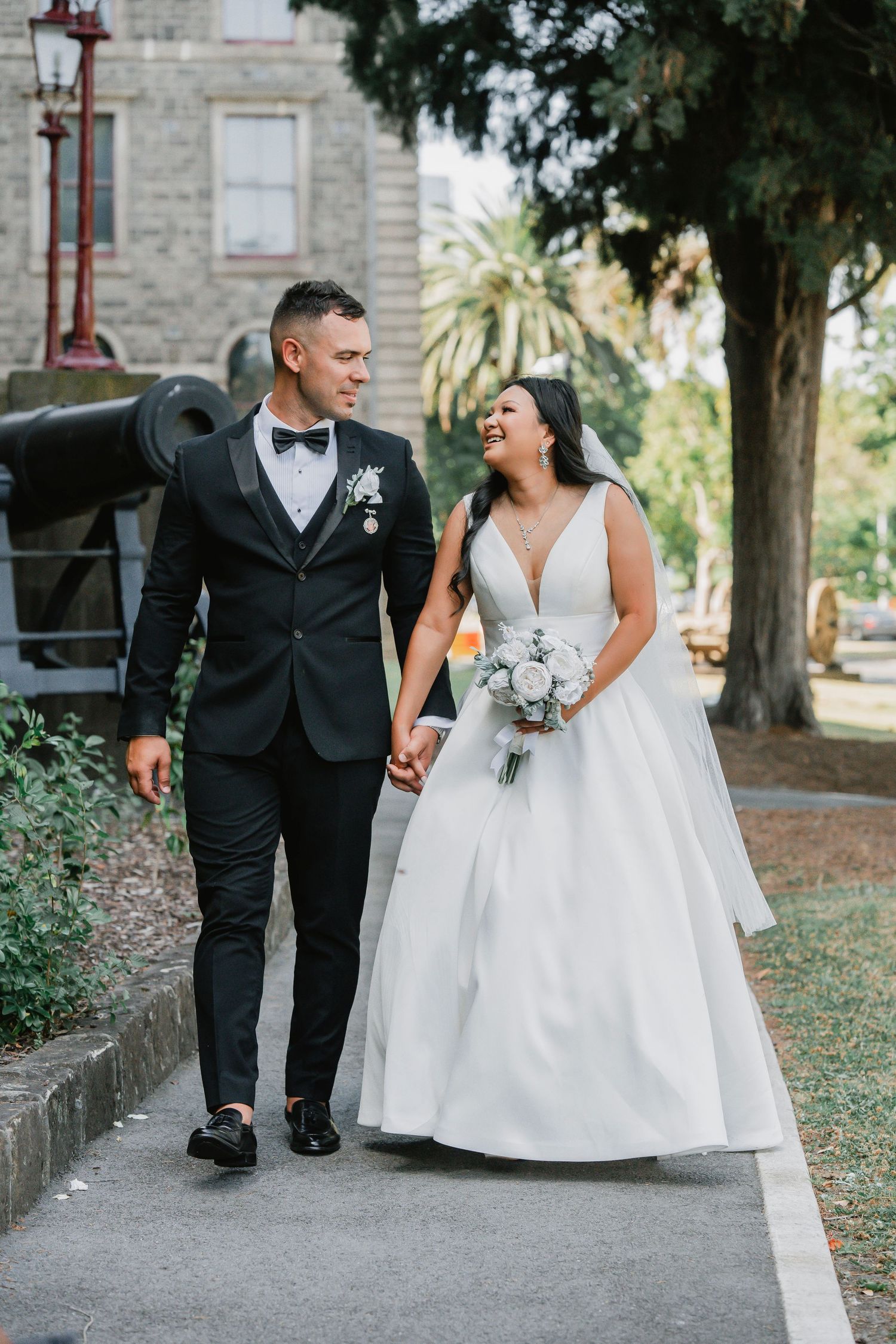 A happy couple in wedding attire walking together on a tree-lined path outside a stone building.