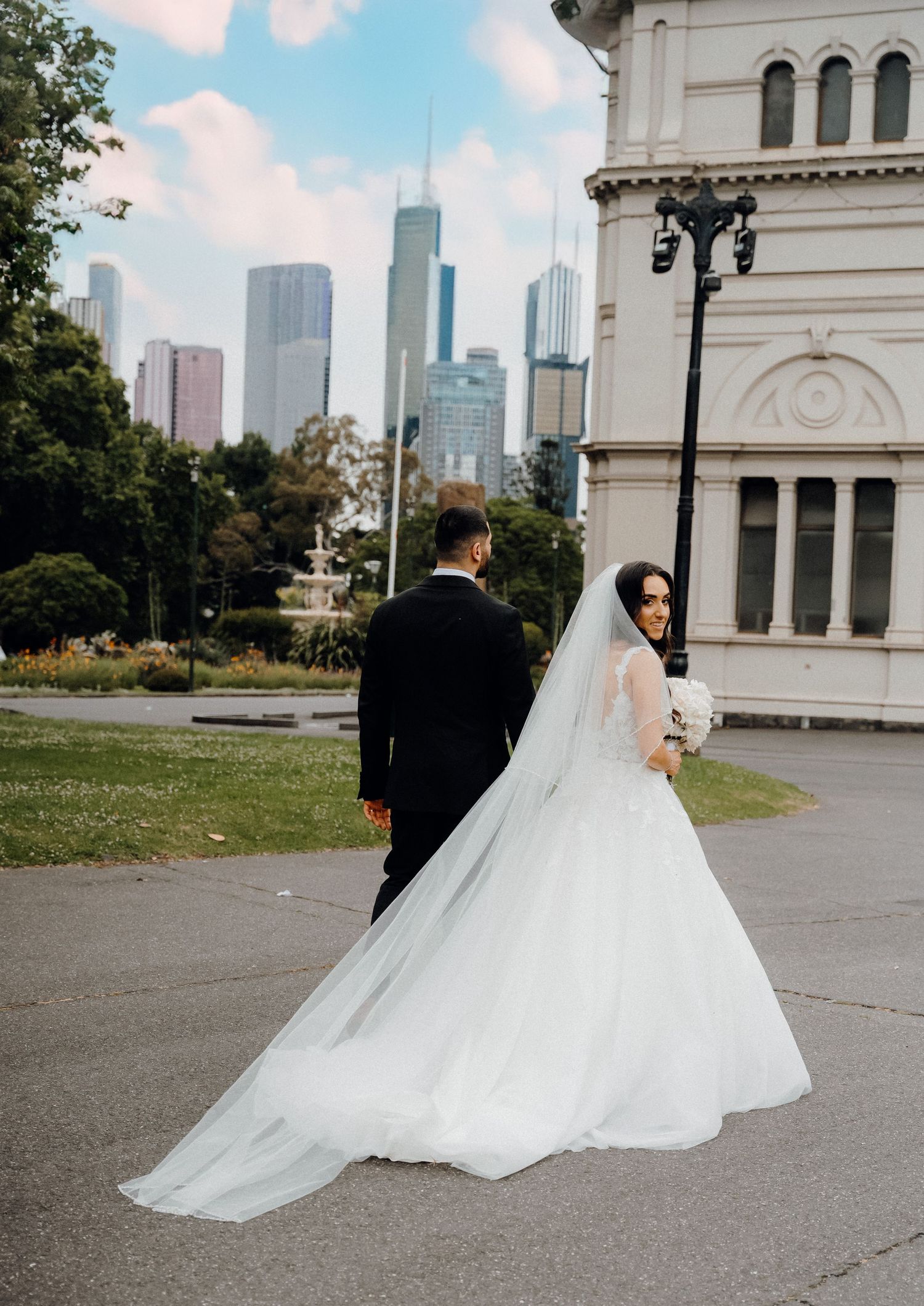 A wedding couple stands against a city backdrop with a historic building and the Chicago skyline visible in the background.