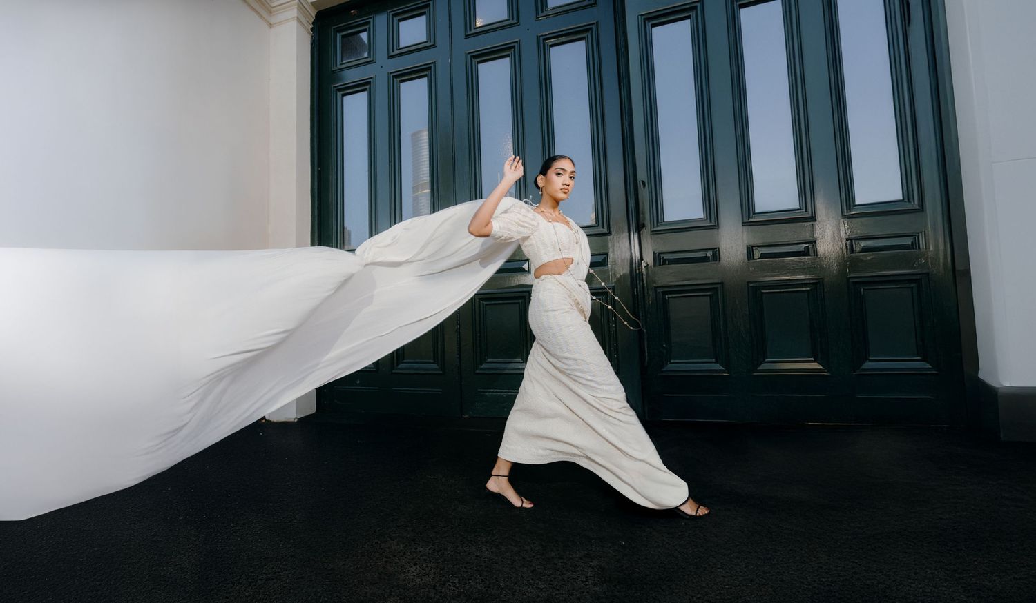 Elegant bride in flowing white dress and veil moves gracefully against dark green doors in architectural setting.