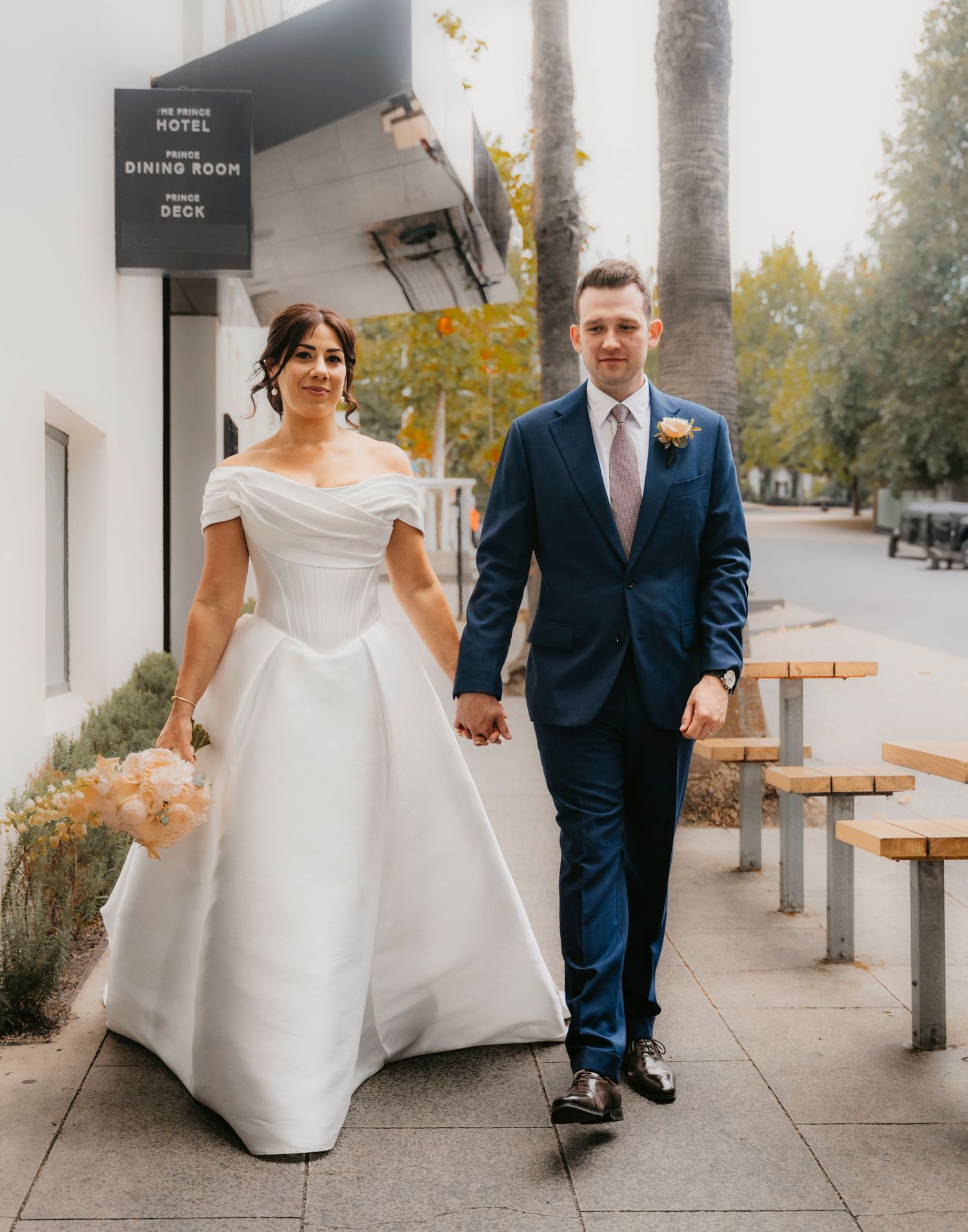 A pair of newlyweds hold hands while walking along a sidewalk with benches and trees, the bride in white and groom in navy.