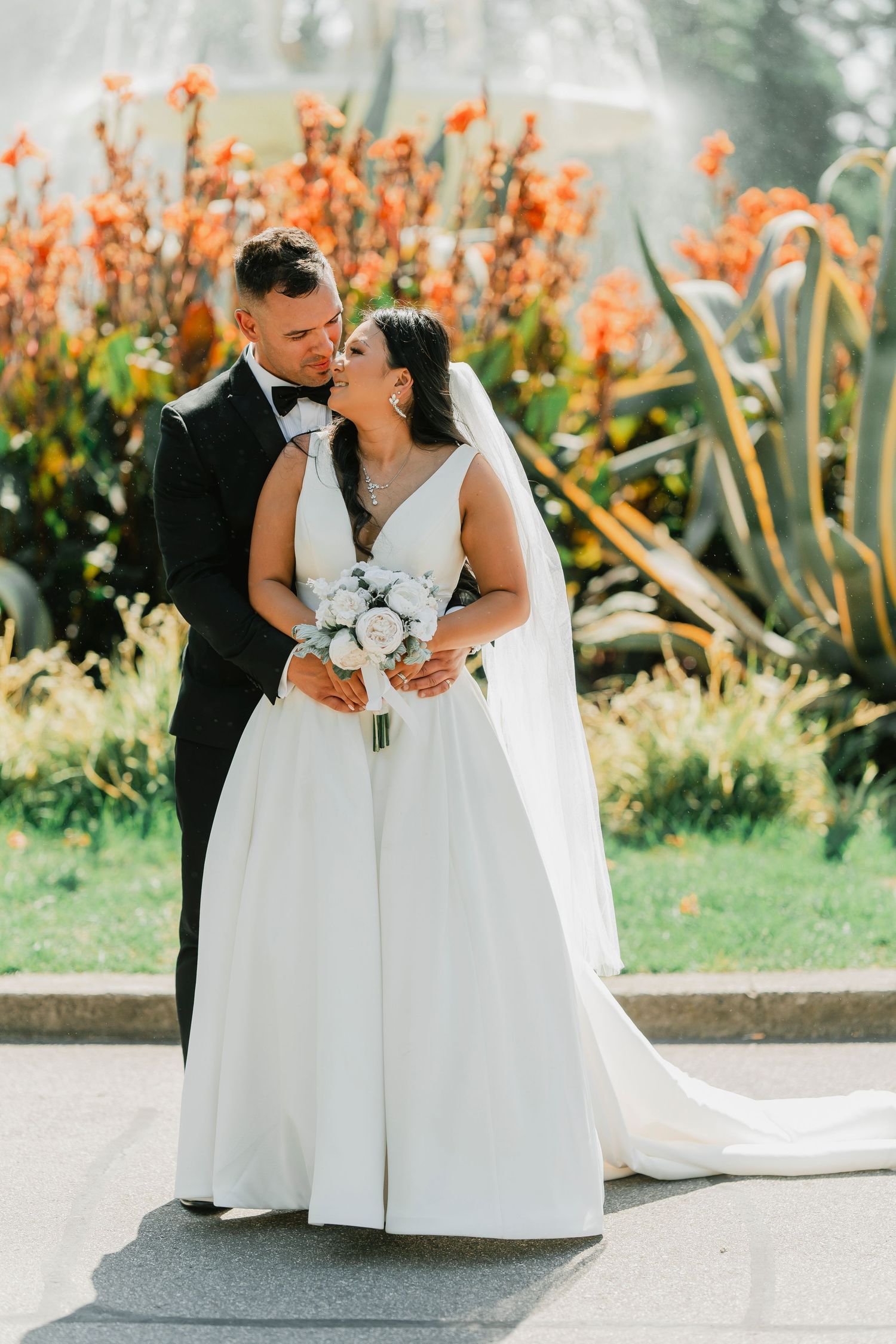 Newlywed couple embraces on garden path with vibrant orange flowers and greenery in background.