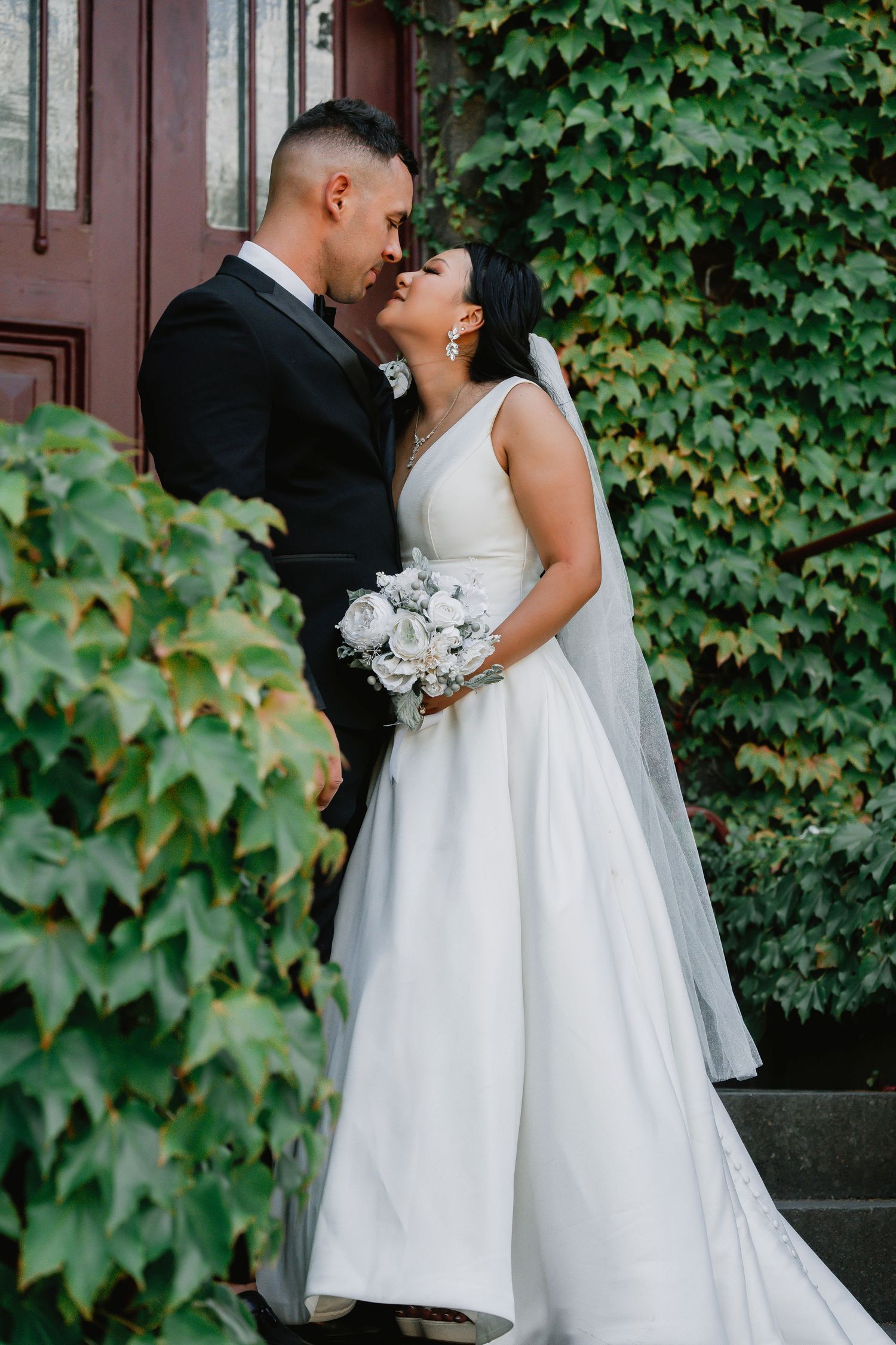 Newlywed couple shares romantic moment against ivy-covered wall, bride holding white bouquet in elegant wedding gown.