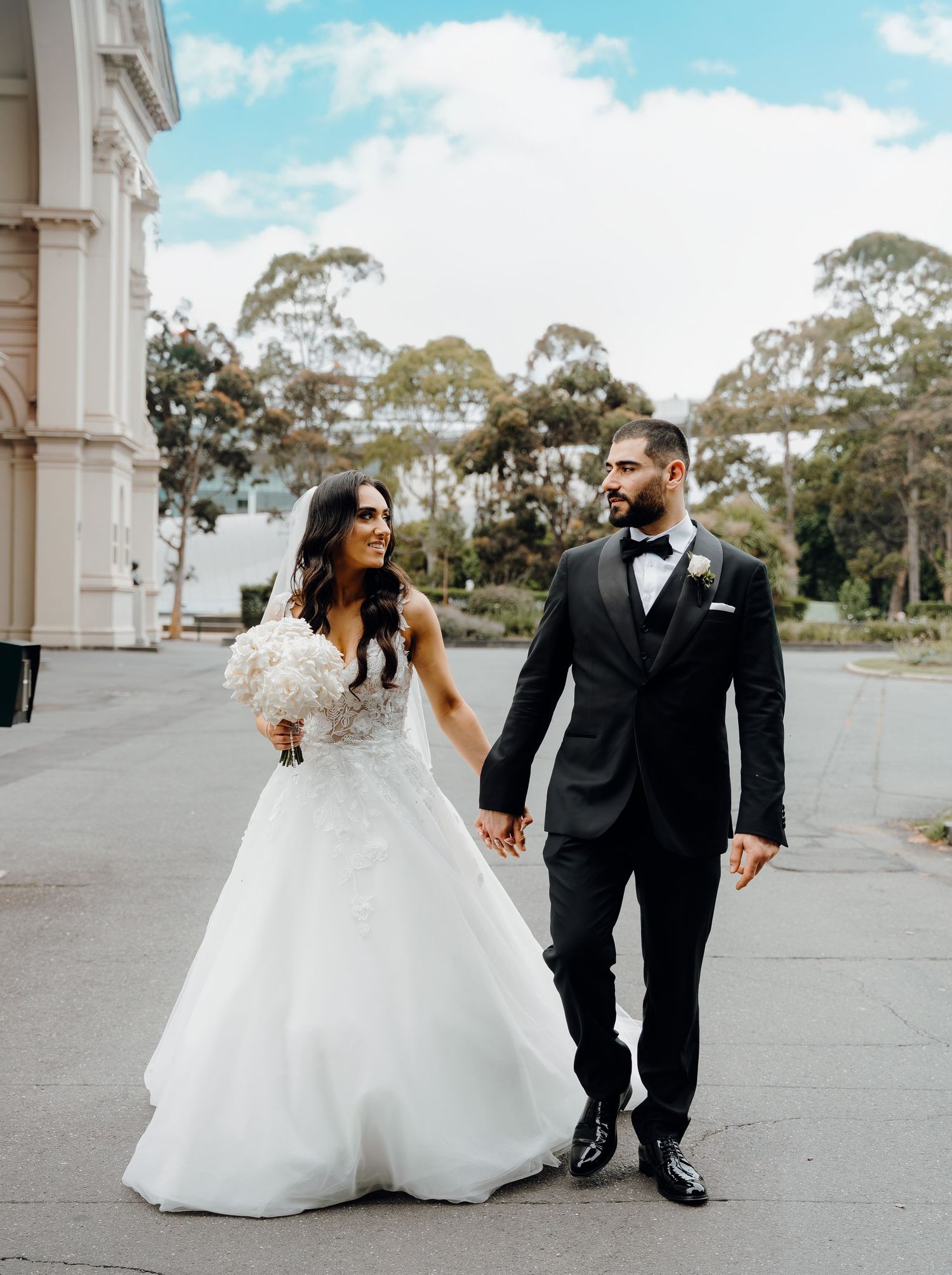 A couple in formal wedding attire walks together outdoors with elegant architecture and trees in the background.