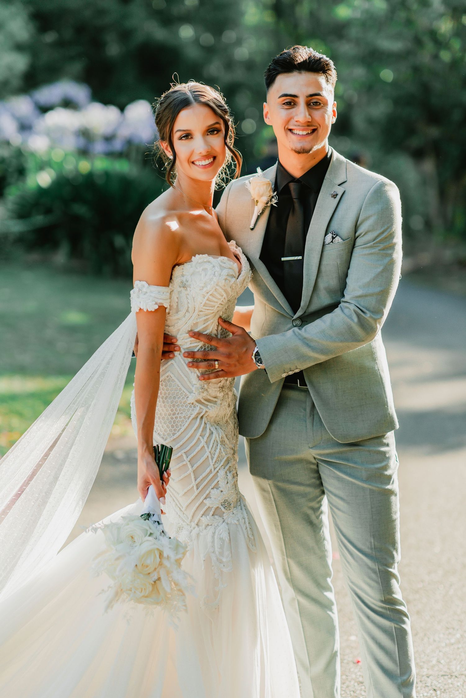 A bride in a fitted lace gown and groom in light gray suit pose together outdoors during their wedding.